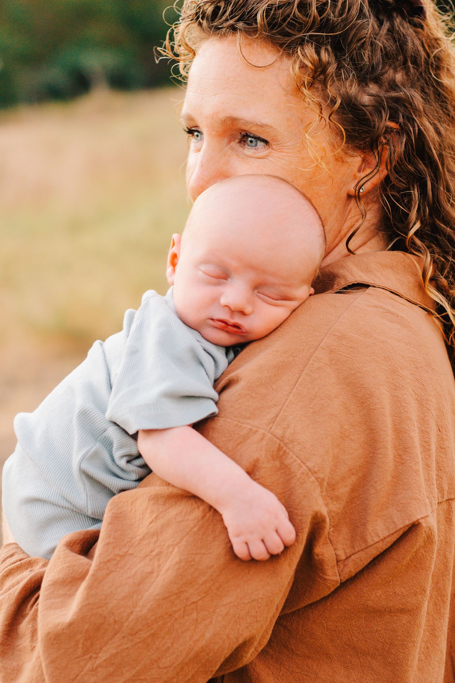 Baby in light blue outfit being held against brown sweater in outdoor setting.