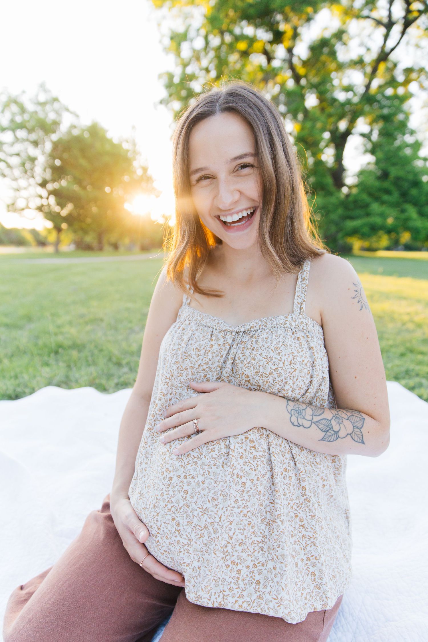 Smiling pregnant woman in floral top holding her baby bump while sitting outdoors in golden sunlight.
