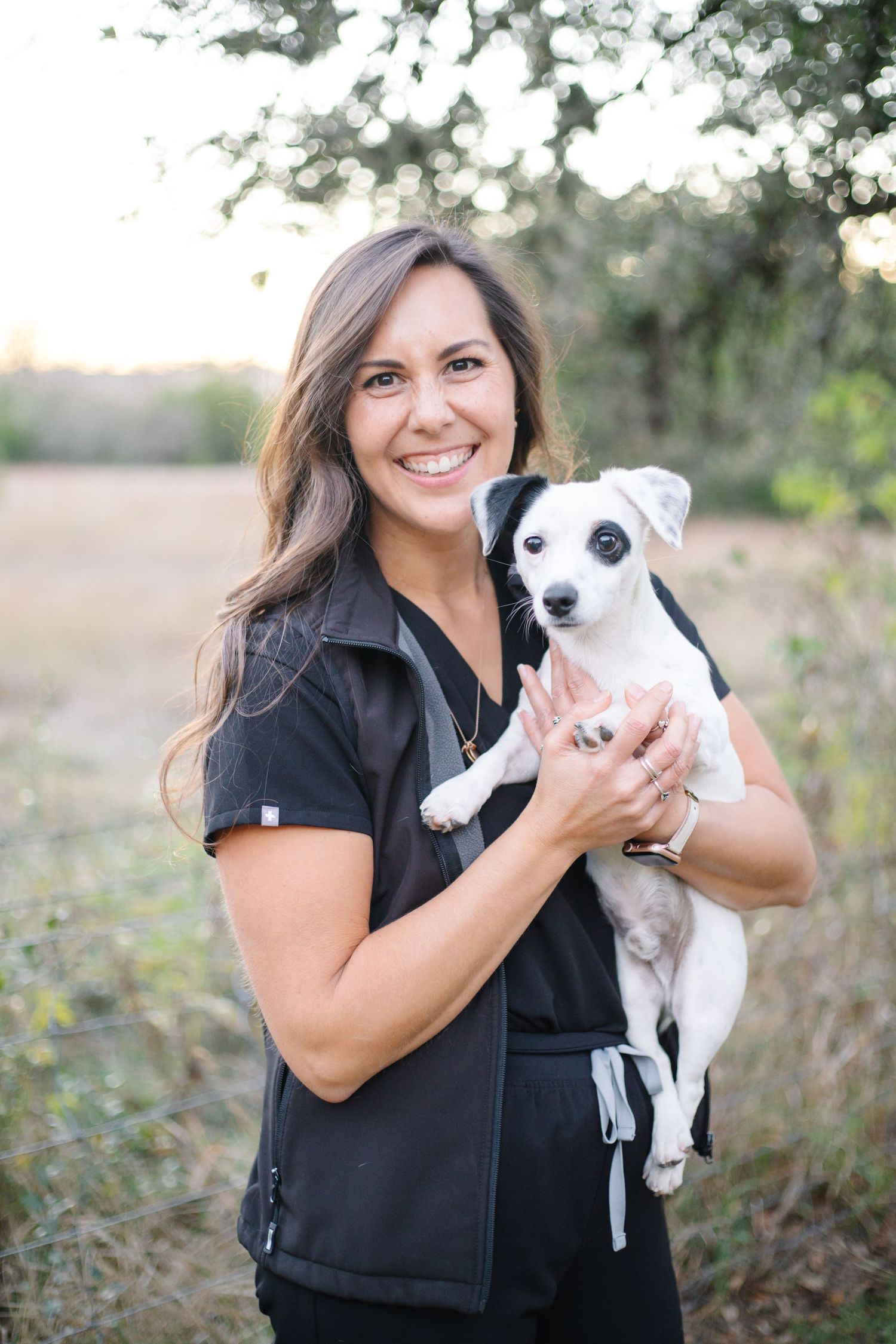Person in black shirt holding small white puppy outdoors during sunset photoshoot.