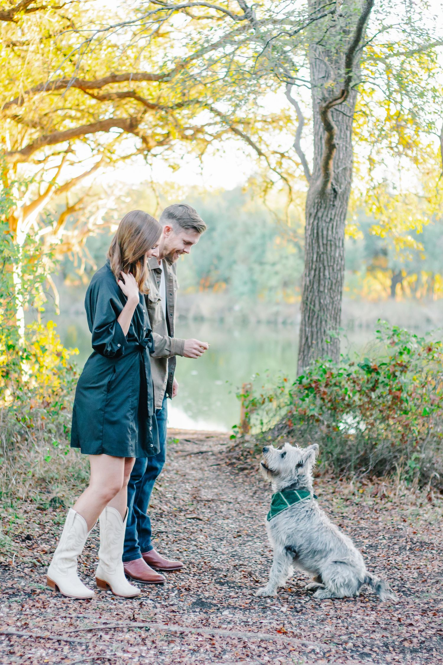 A couple plays with their small gray dog on a leaf-covered trail during golden hour.
