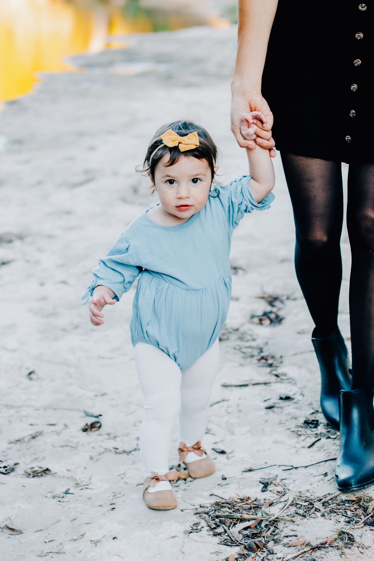 Small child in light blue dress taking first steps on beach while holding parent's hand.