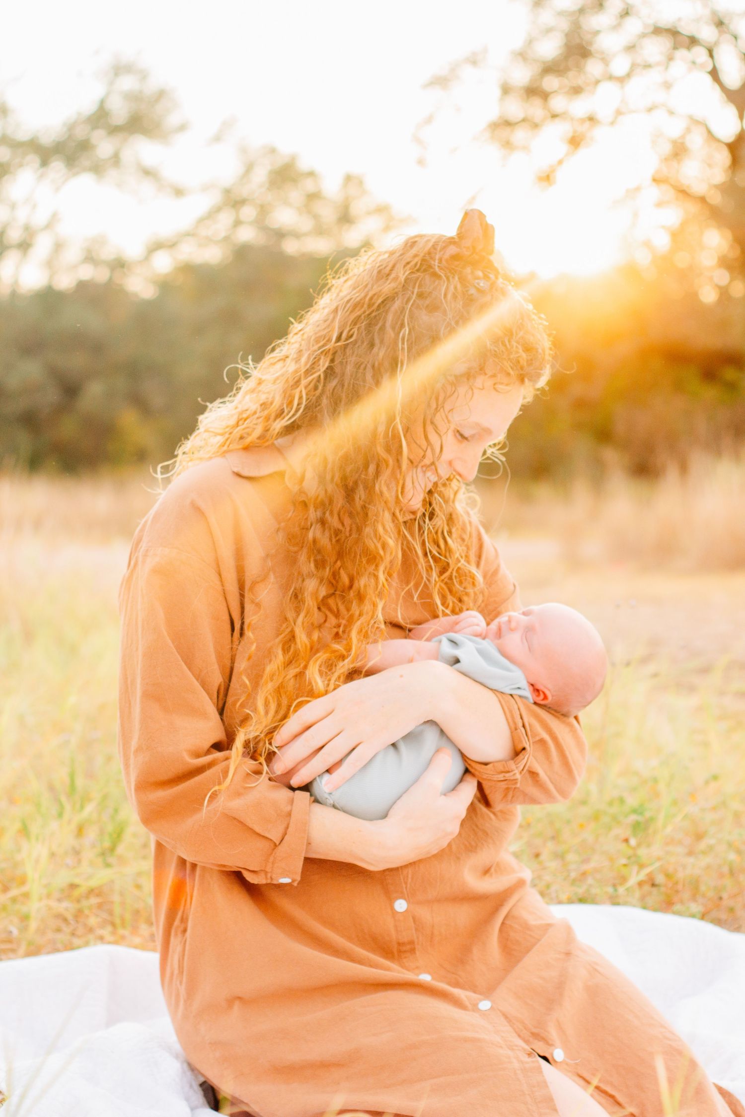 Parent in orange sweater holds and embraces baby against golden sunset backdrop in outdoor setting.