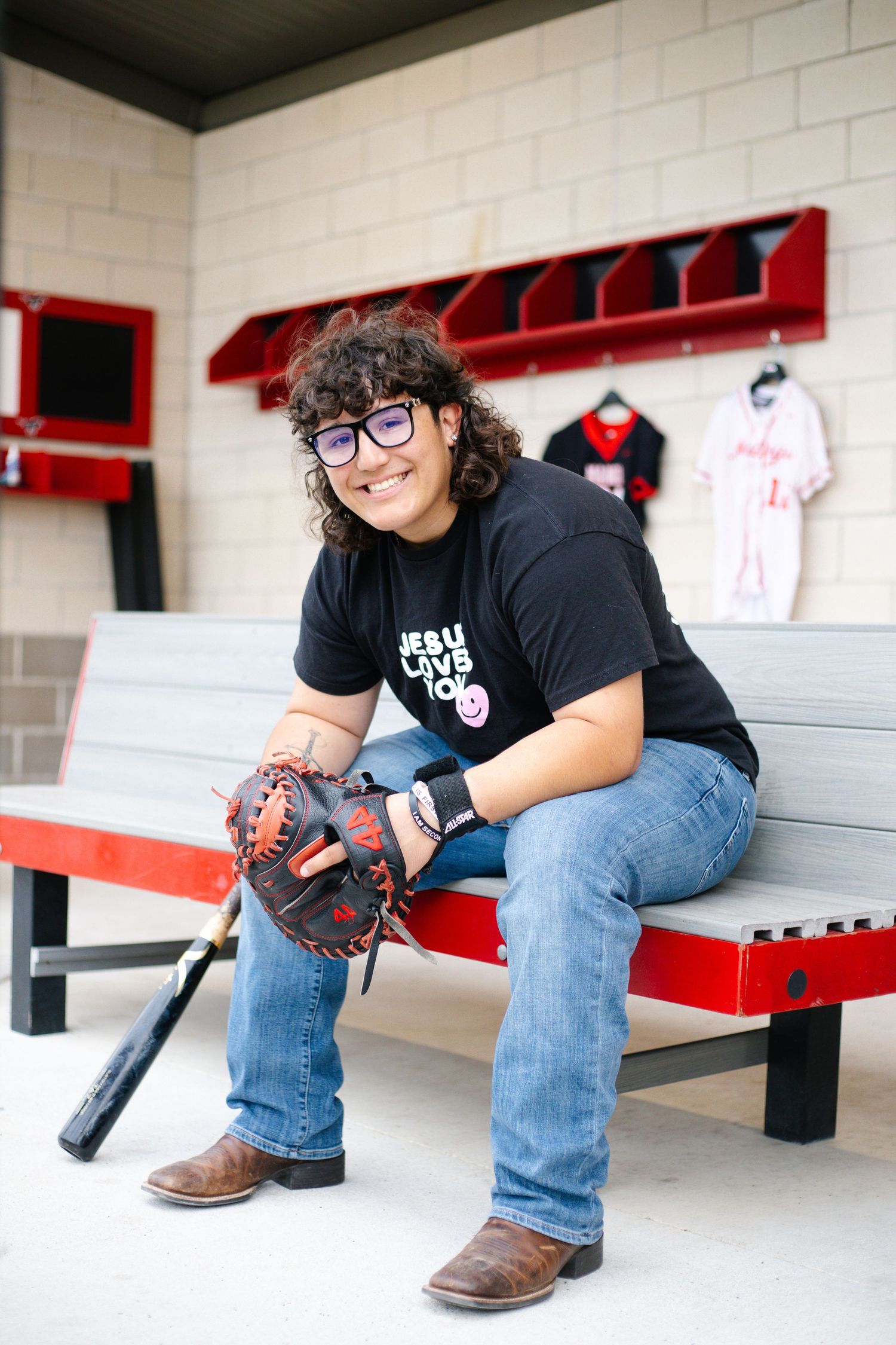 Smiling student sits on dugout bench holding baseball glove and bat wearing Jesus Loves You shirt and jeans.
