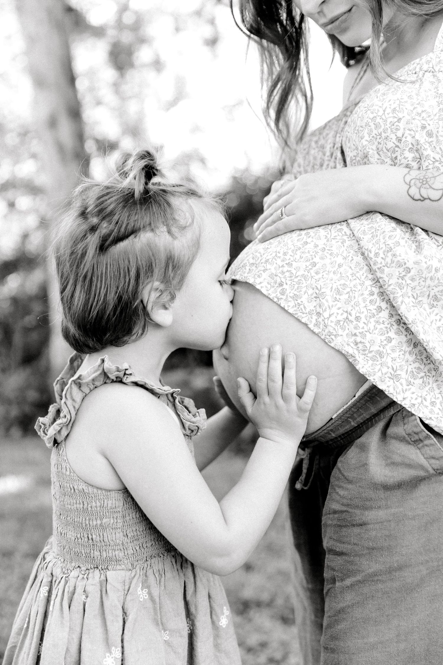 Black and white photo of a young girl kissing her pregnant mother's baby bump outdoors.