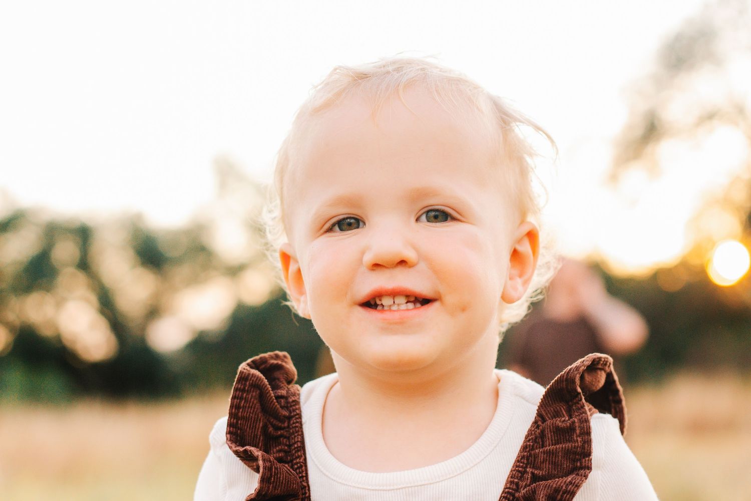 Baby wearing brown ruffled outfit smiles brightly during outdoor photo session.