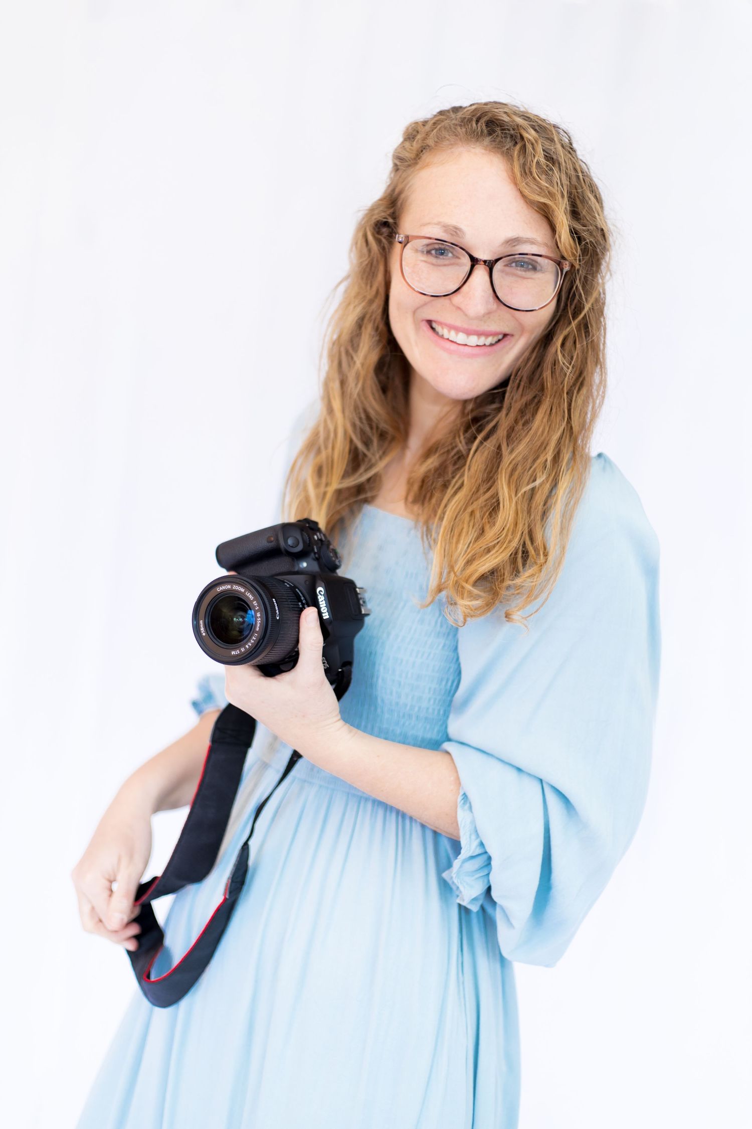 Professional photographer in light blue dress holding camera and smiling against white background.