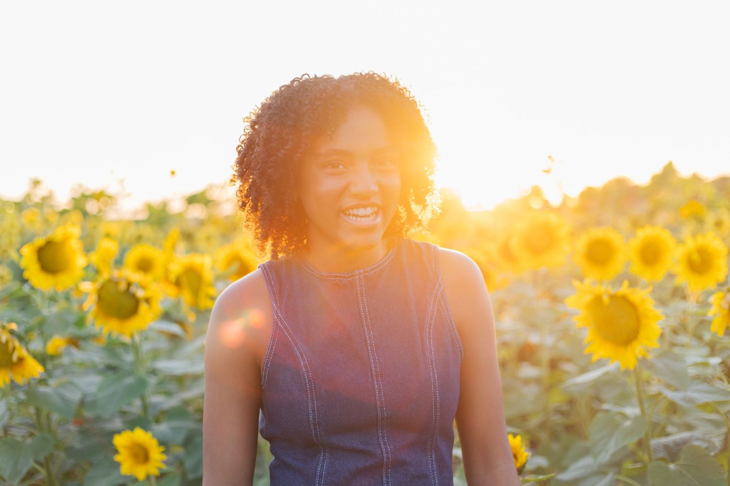 Smiling young Black woman standing in a vibrant sunflower field bathed in golden sunset light.