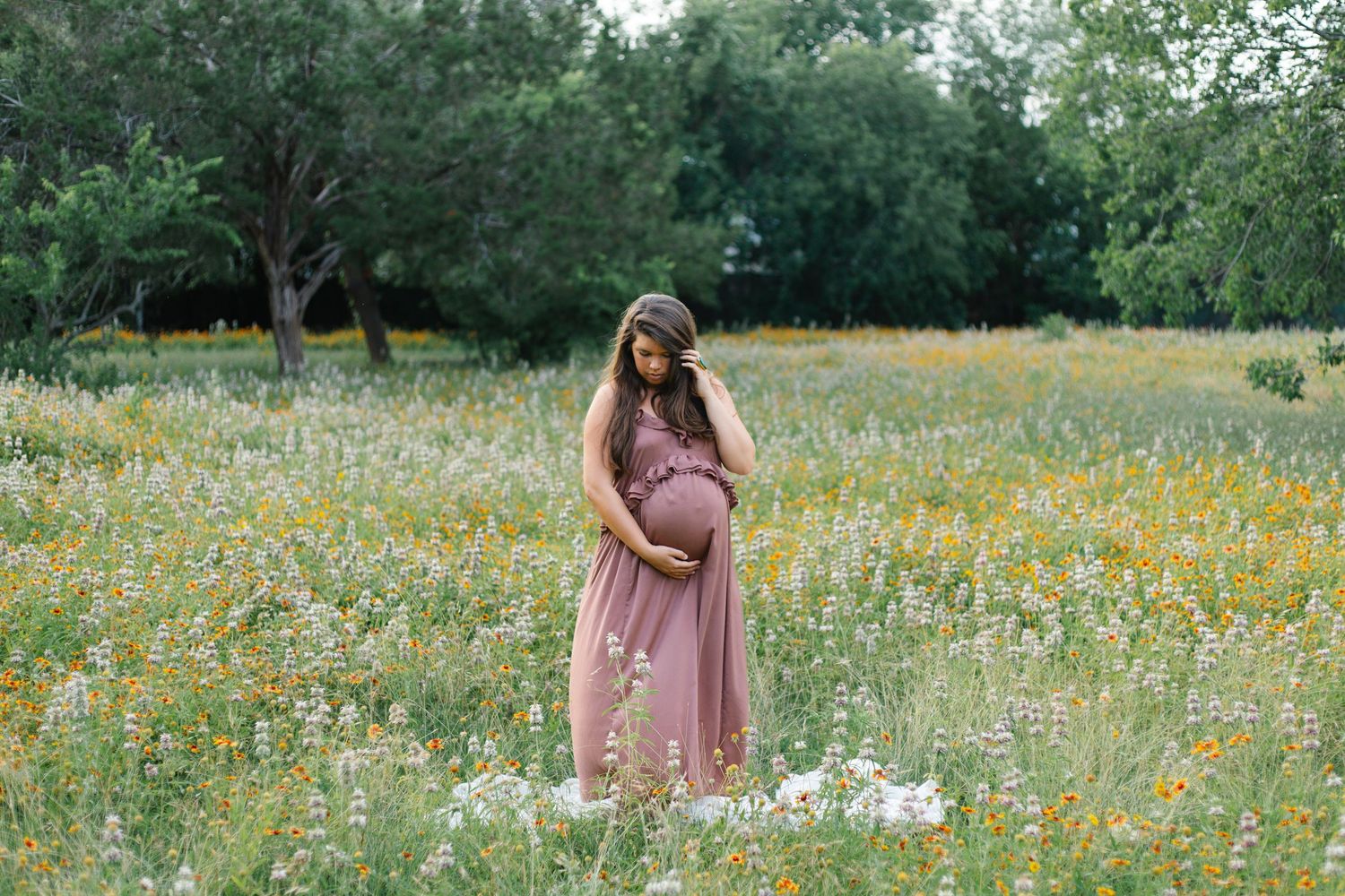 Pregnant woman in pink maxi dress standing in wildflower meadow, cradling baby bump surrounded by colorful blooms and trees.