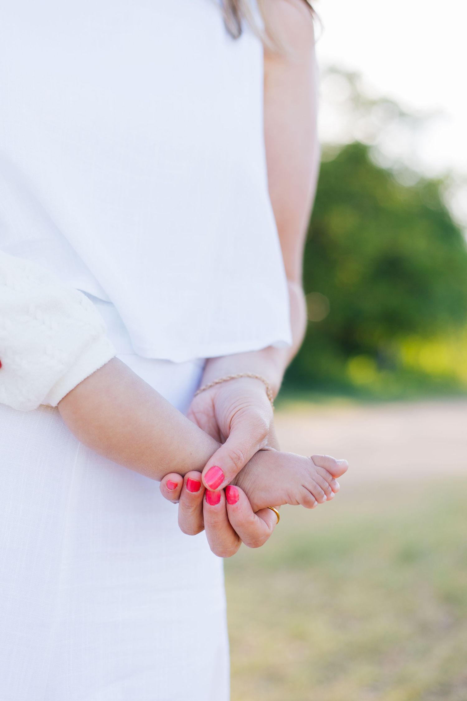 A woman in white dress holds a baby's tiny foot with pink manicured nails outdoors.