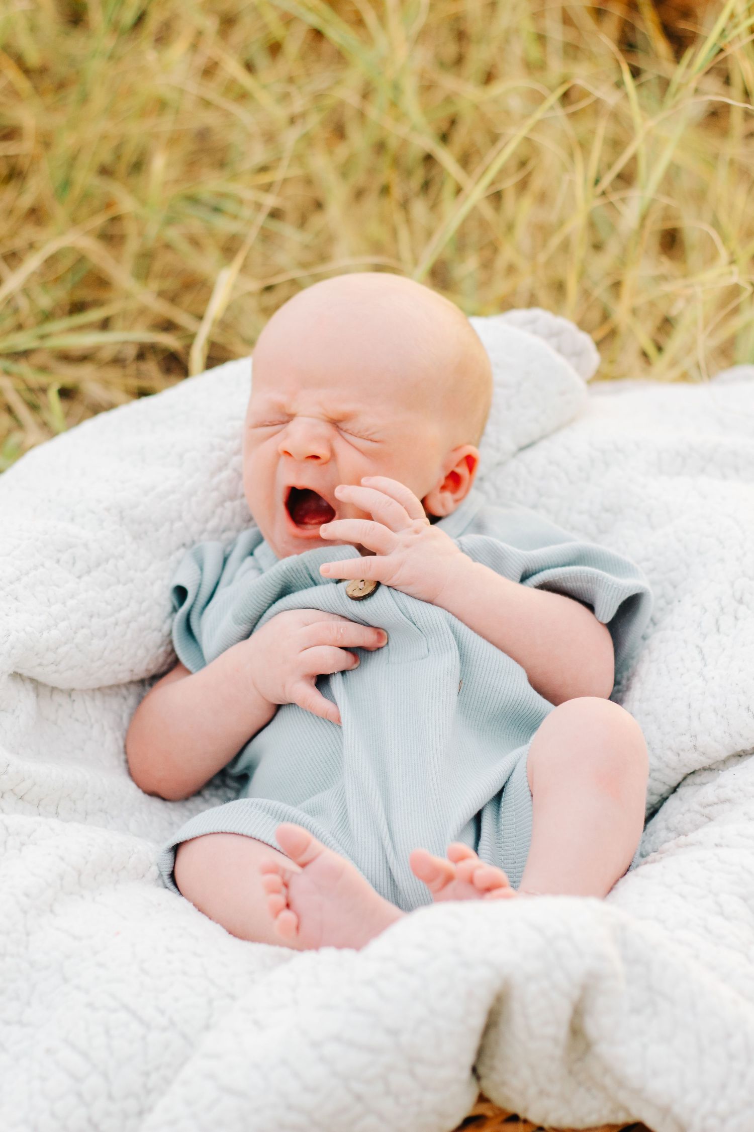 Baby in light blue outfit yawning while lying on white blanket outdoors.