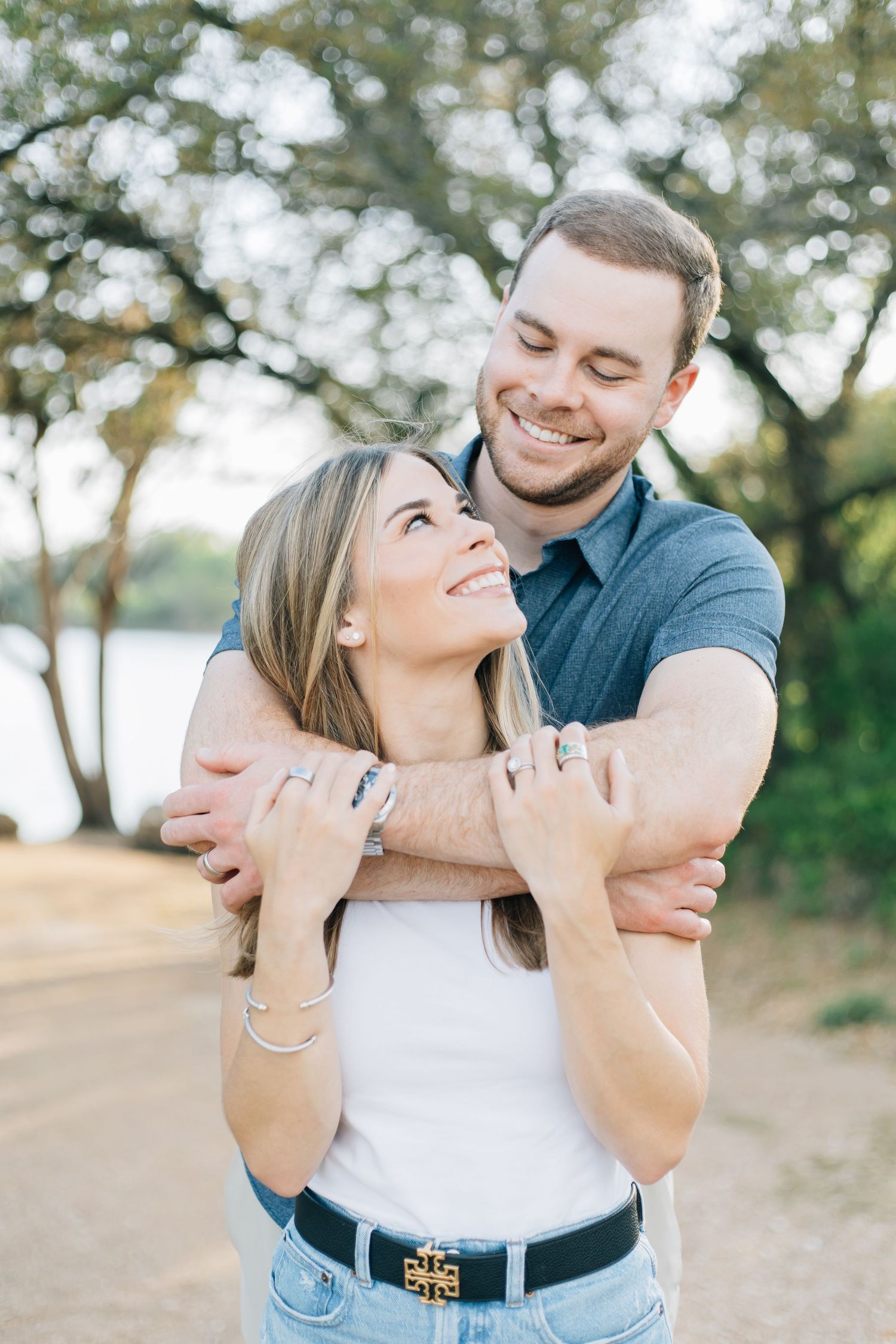Happy couple embracing outdoors near a lake, woman smiling up at man hugging her.