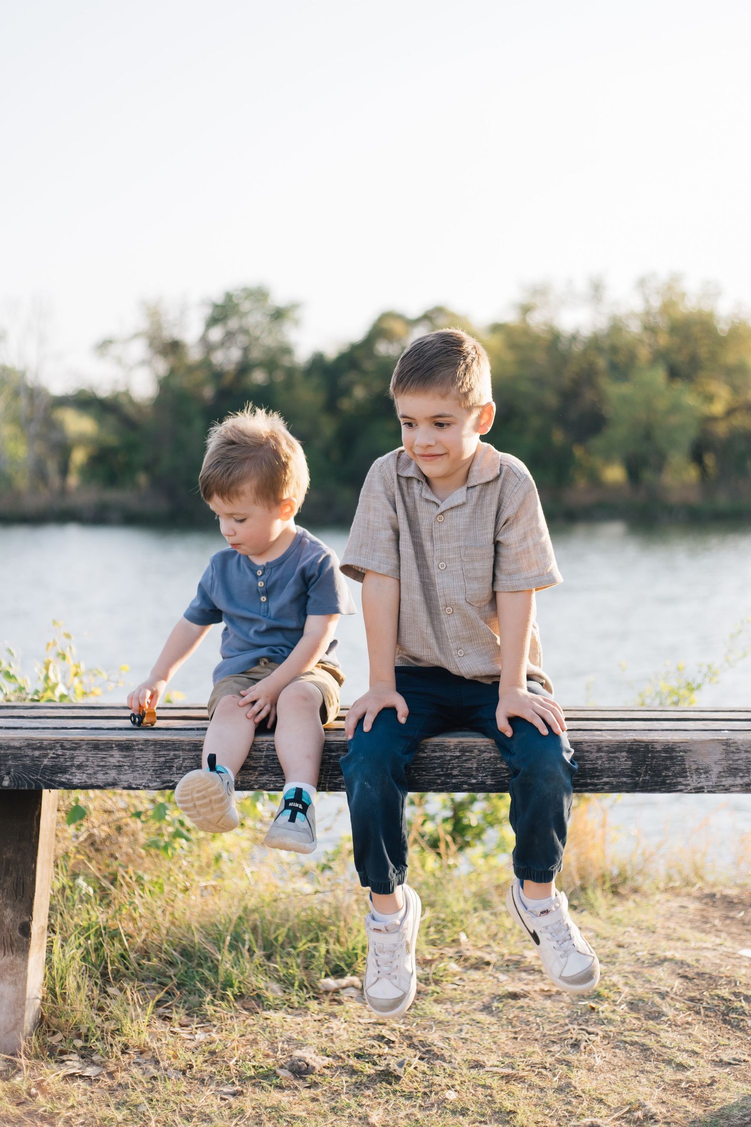 Two young brothers sitting on a wooden bench near a lake on a sunny day.