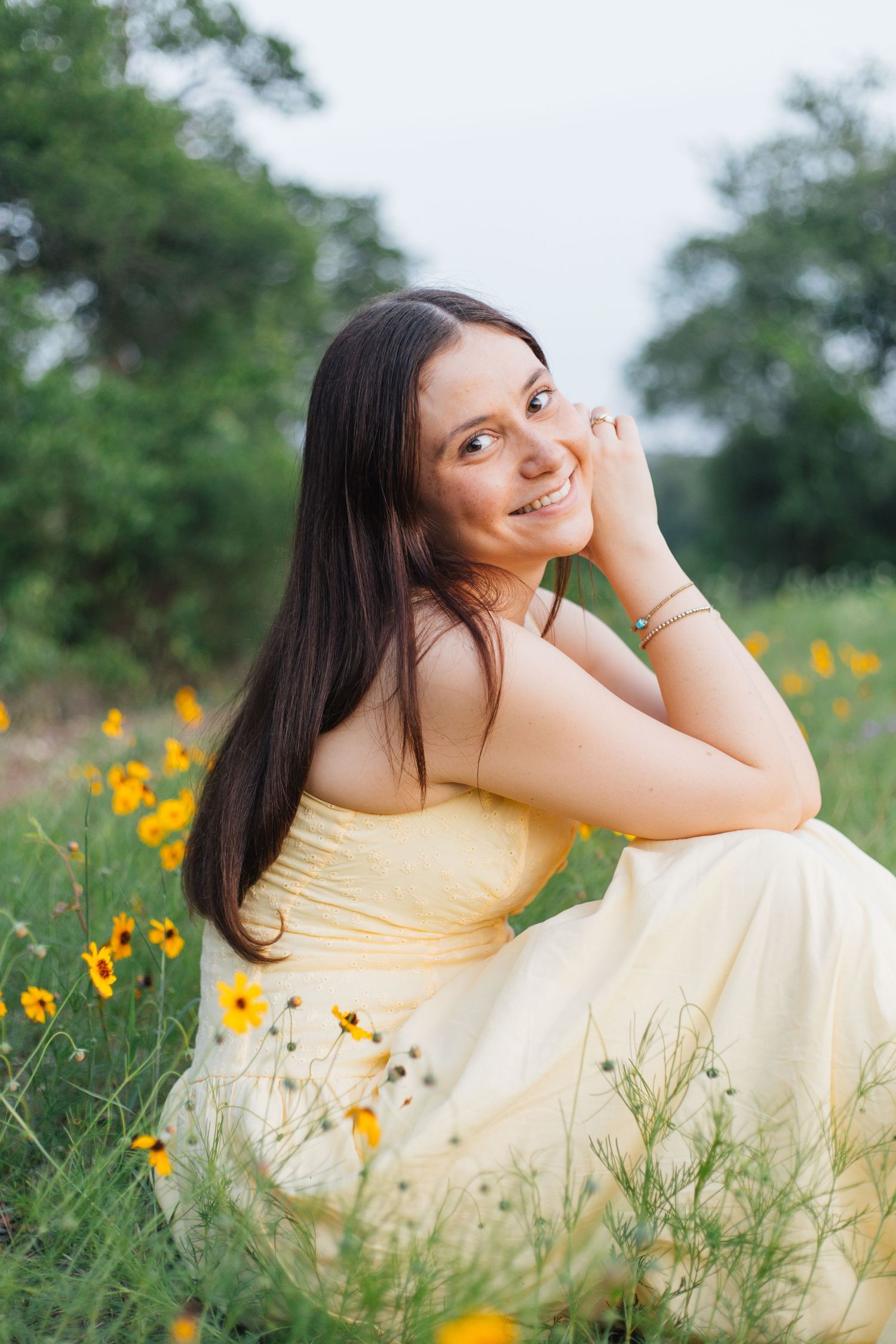 Smiling brunette woman in yellow dress sitting among yellow wildflowers in green field. Senior Portraits Round Rock TX