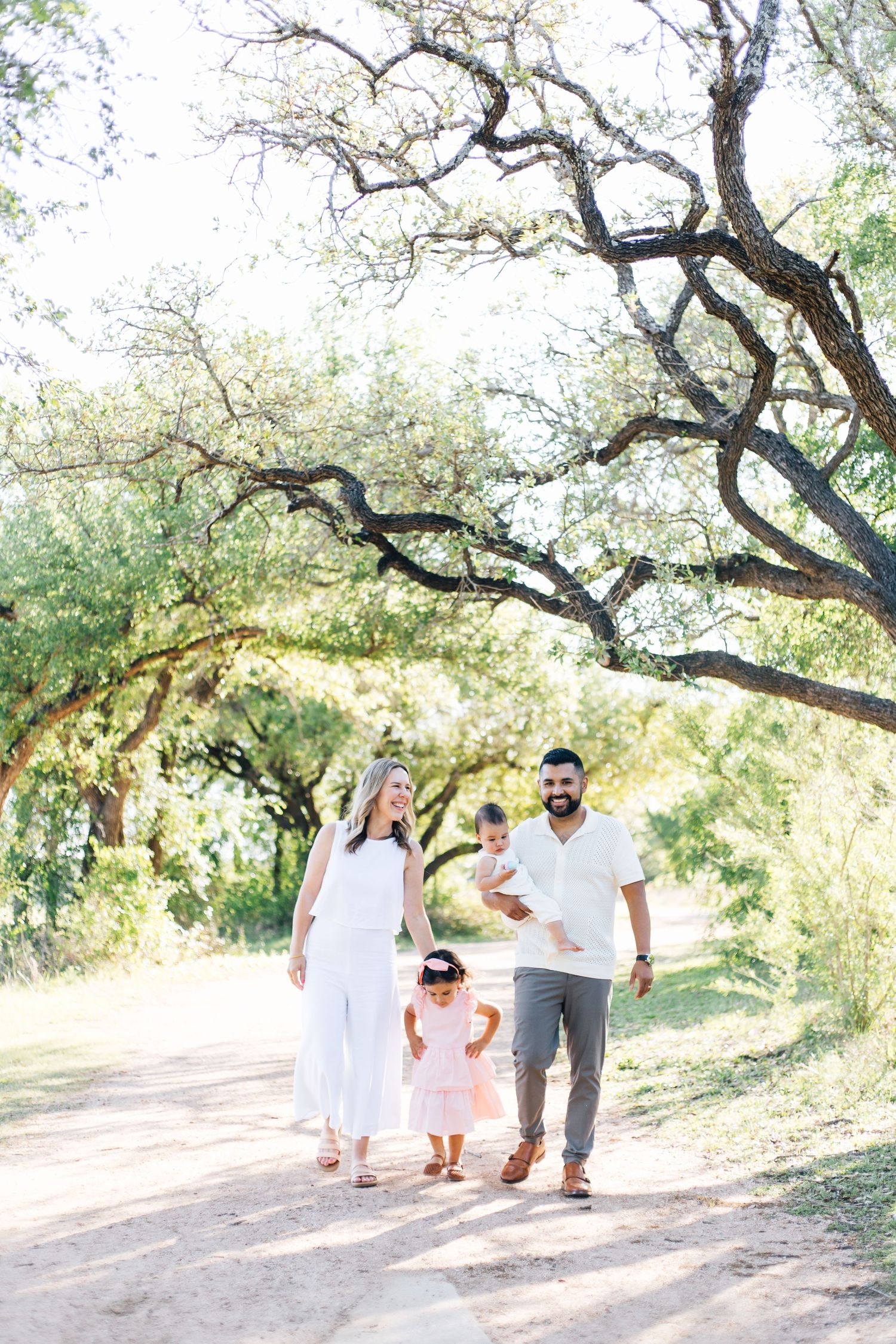 Family of four walking on a sunny dirt path under large oak trees, dressed in white and neutral tones.