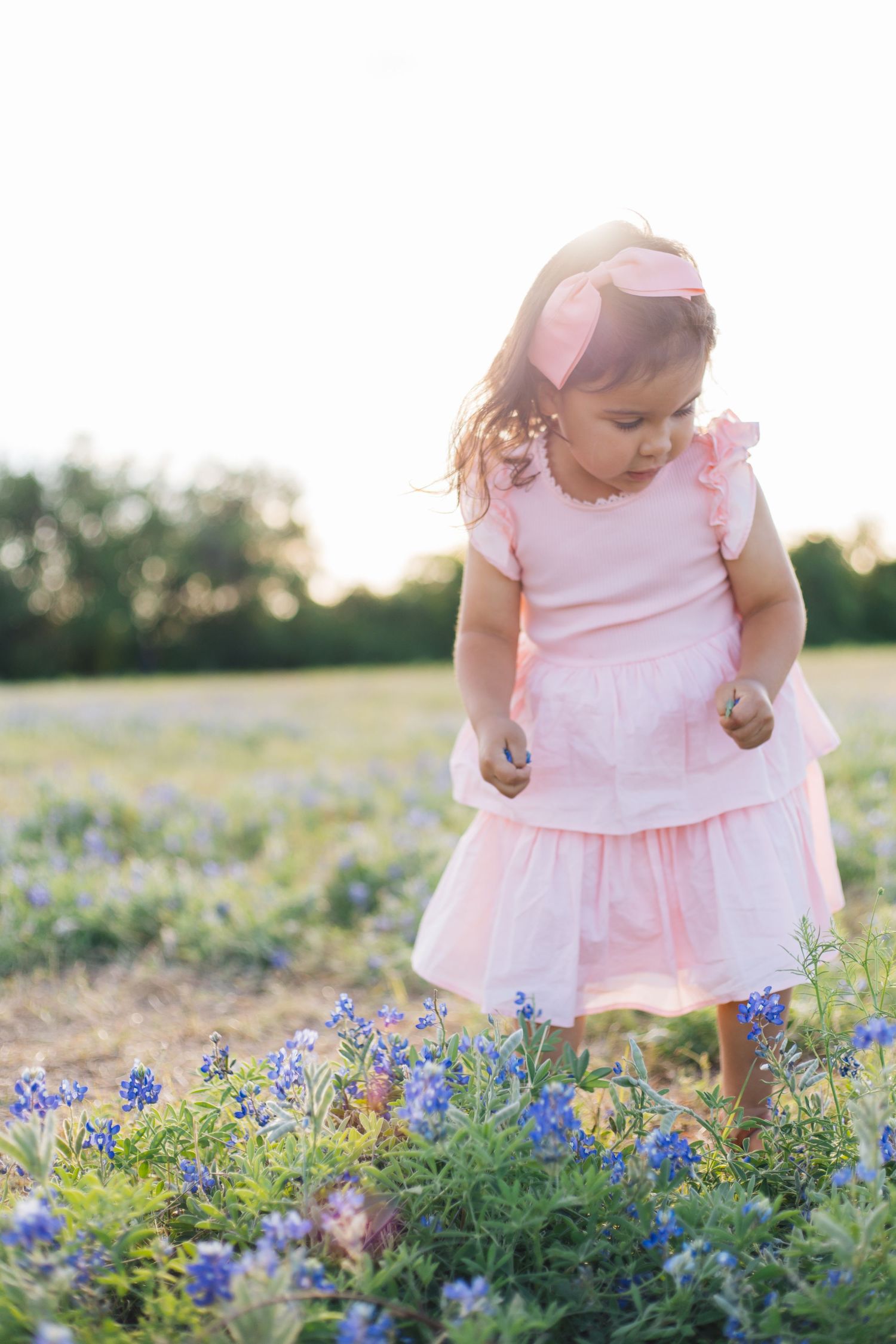 Toddler girl in pink dress and bow headband picking bluebonnets in a sunlit Texas field.