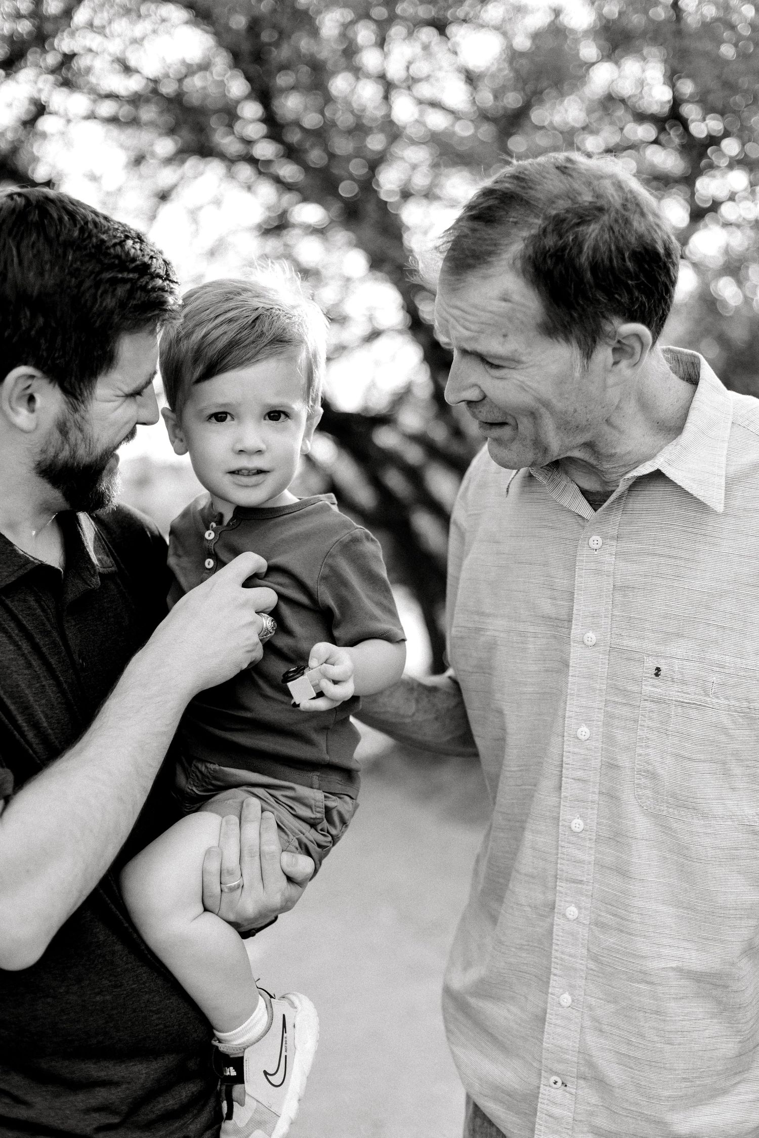 Black and white photo of two men with a toddler boy outdoors among trees.