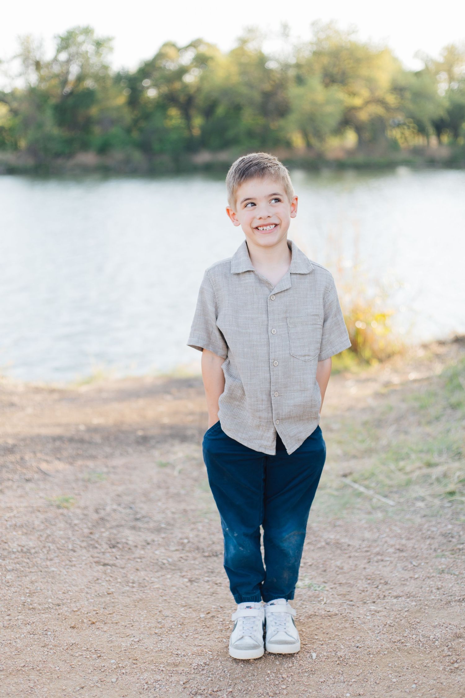 Young boy in gray shirt and jeans smiling outdoors by a lake on a sunny day.