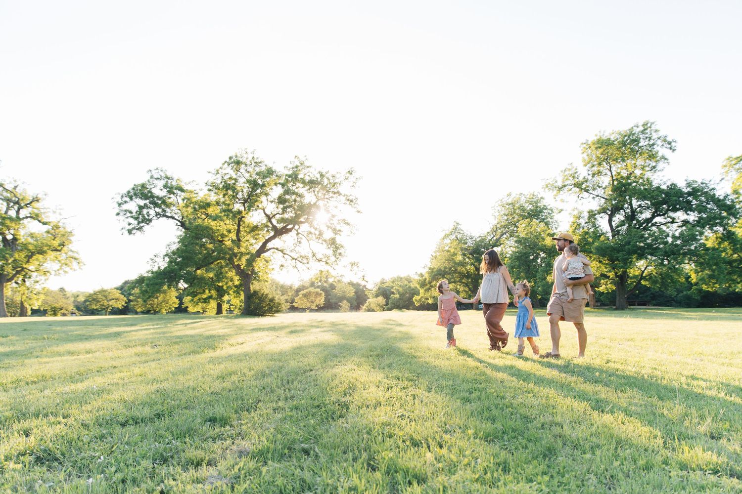 Family of four holding hands walking through a lush green park at golden hour with tall trees in background.