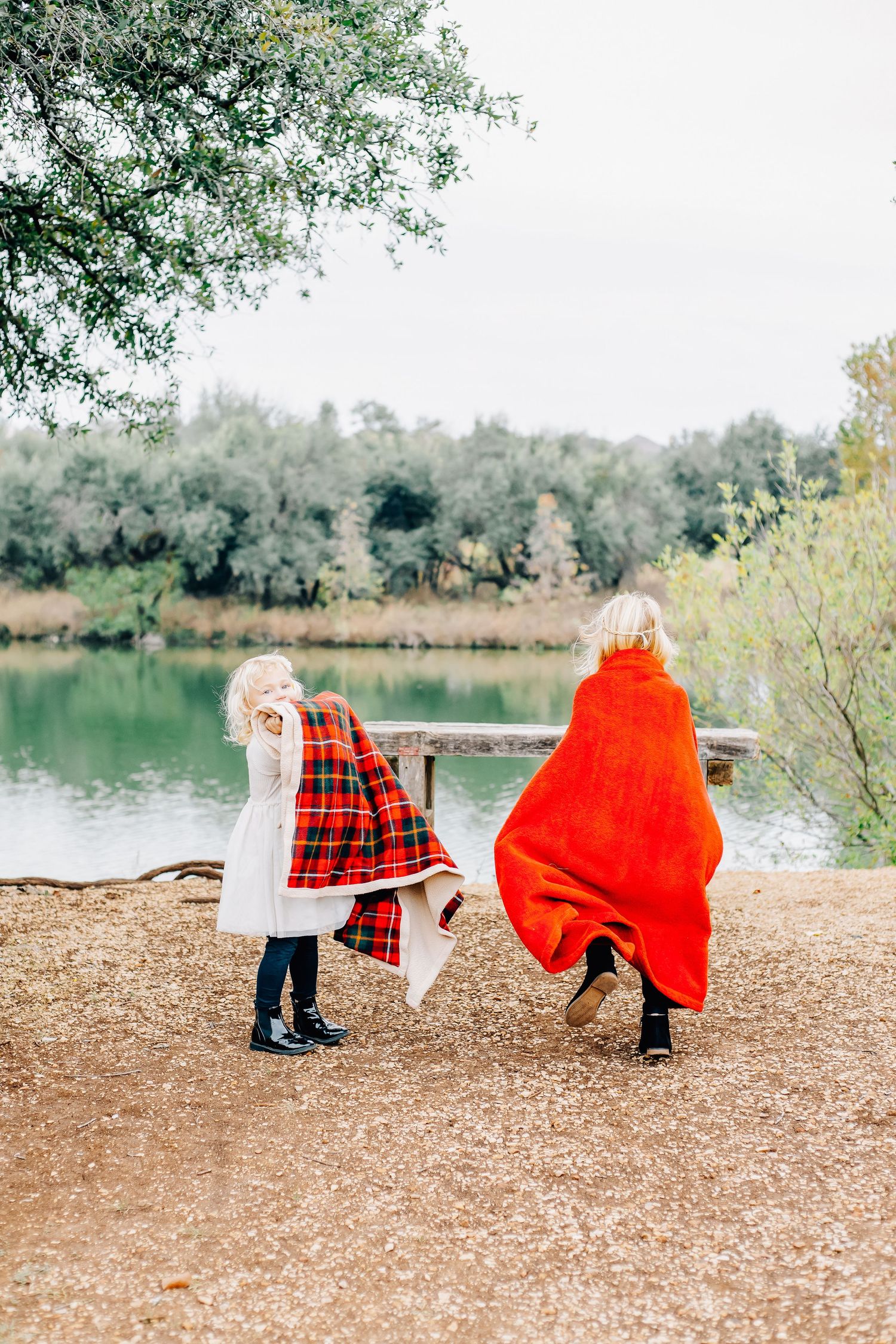 Two people wrapped in red and plaid blankets stand by a serene lake.