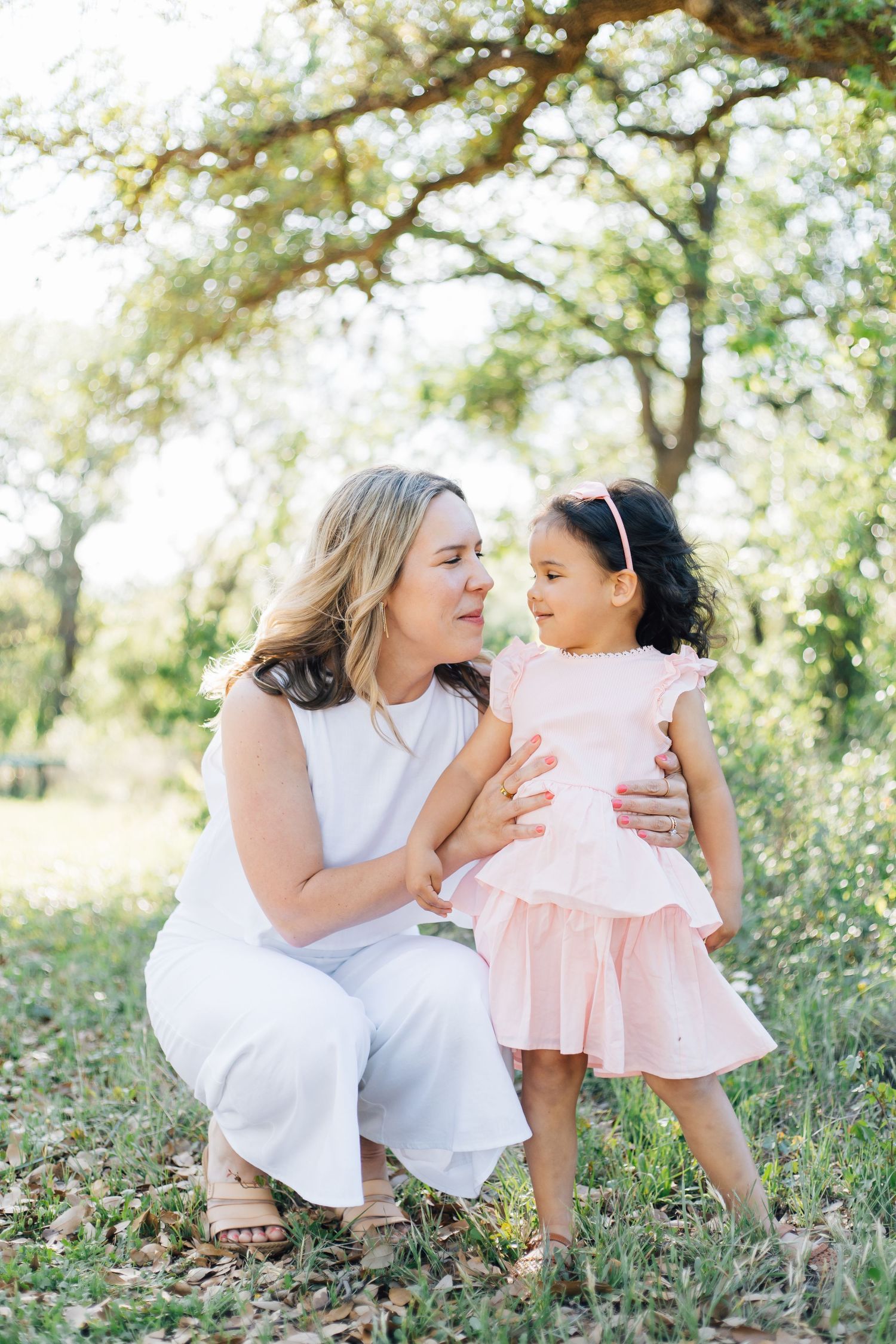 A woman in white crouches beside a toddler girl in a pink dress outdoors among green trees.