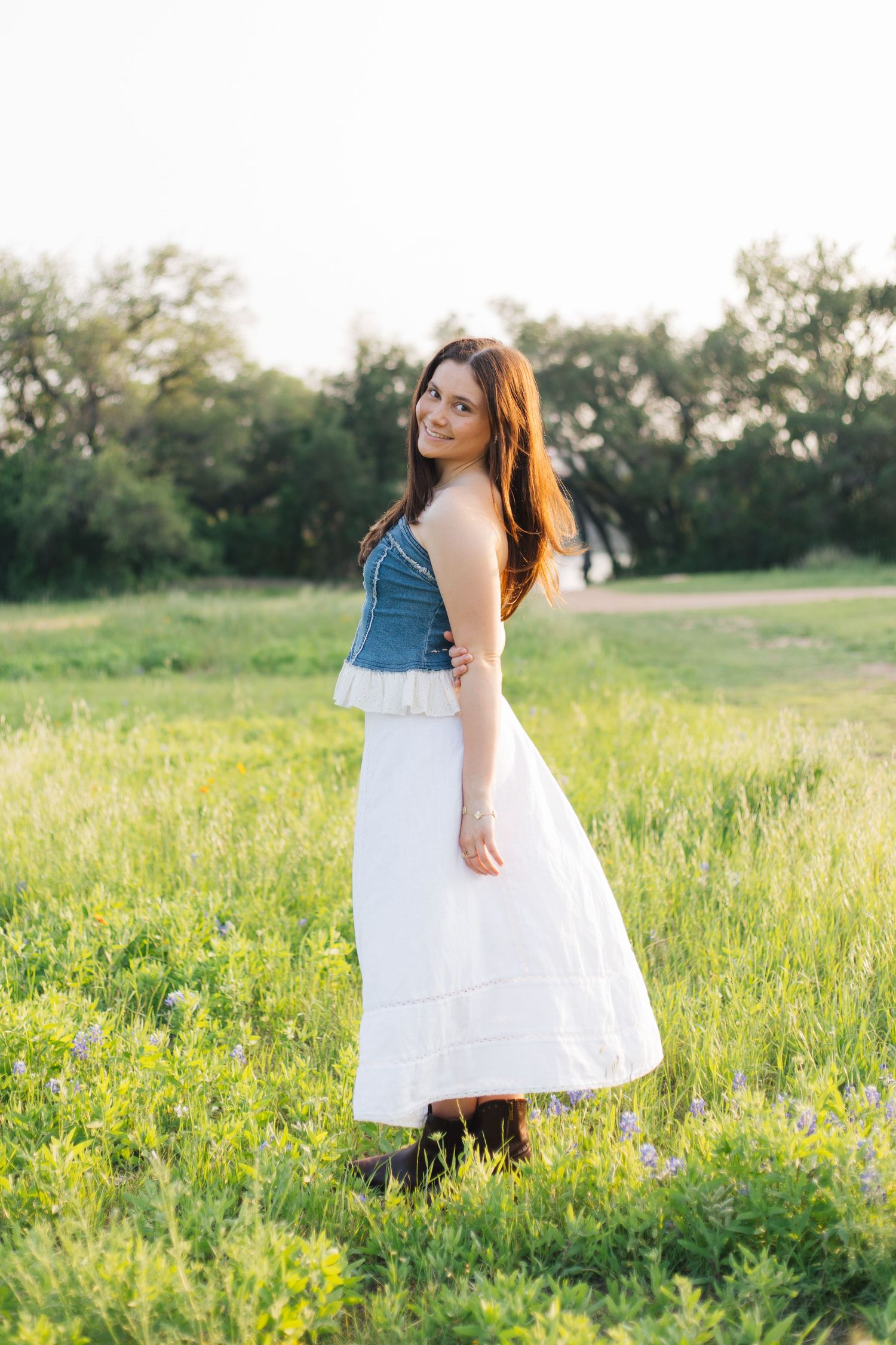 Young woman in denim tube top and white maxi skirt standing in a sunny green meadow. Senior Portraits Round Rock TX