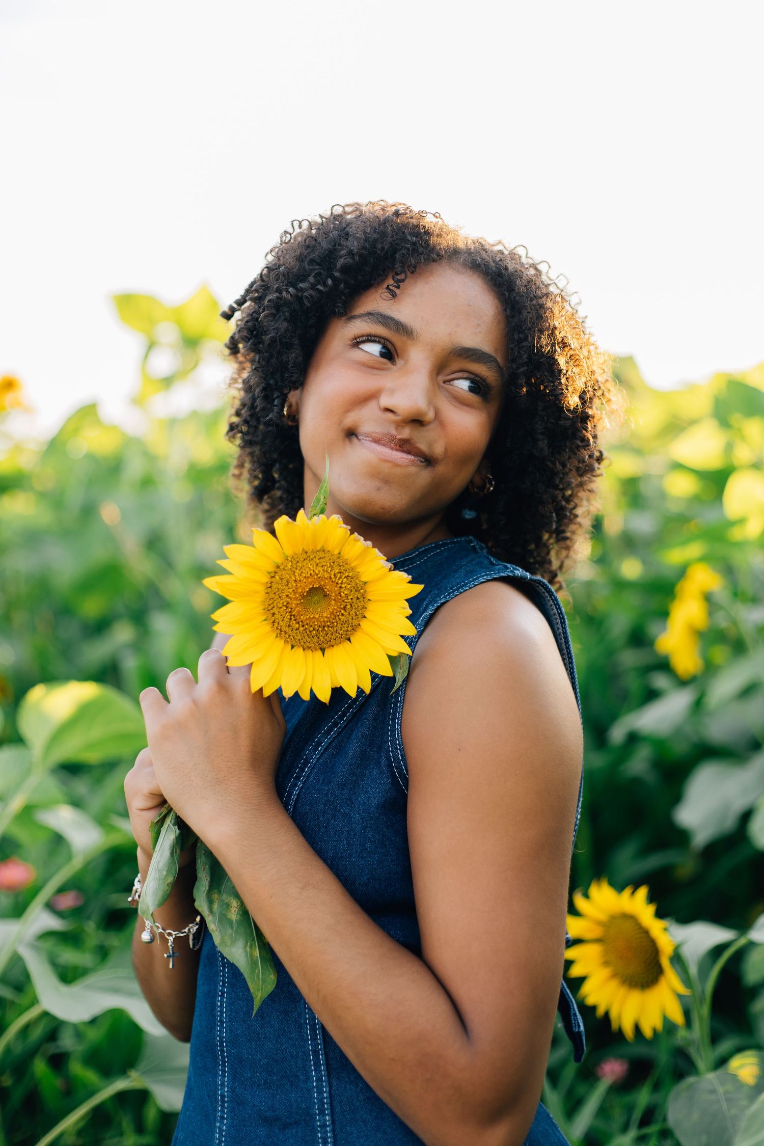 Young woman with curly hair holding a bright yellow sunflower in a blooming sunflower field, wearing a denim dress.