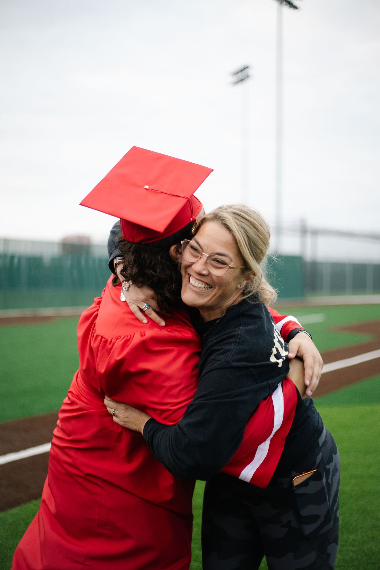 A graduate in red cap and gown shares a joyful hug with a smiling woman on a sports field.
