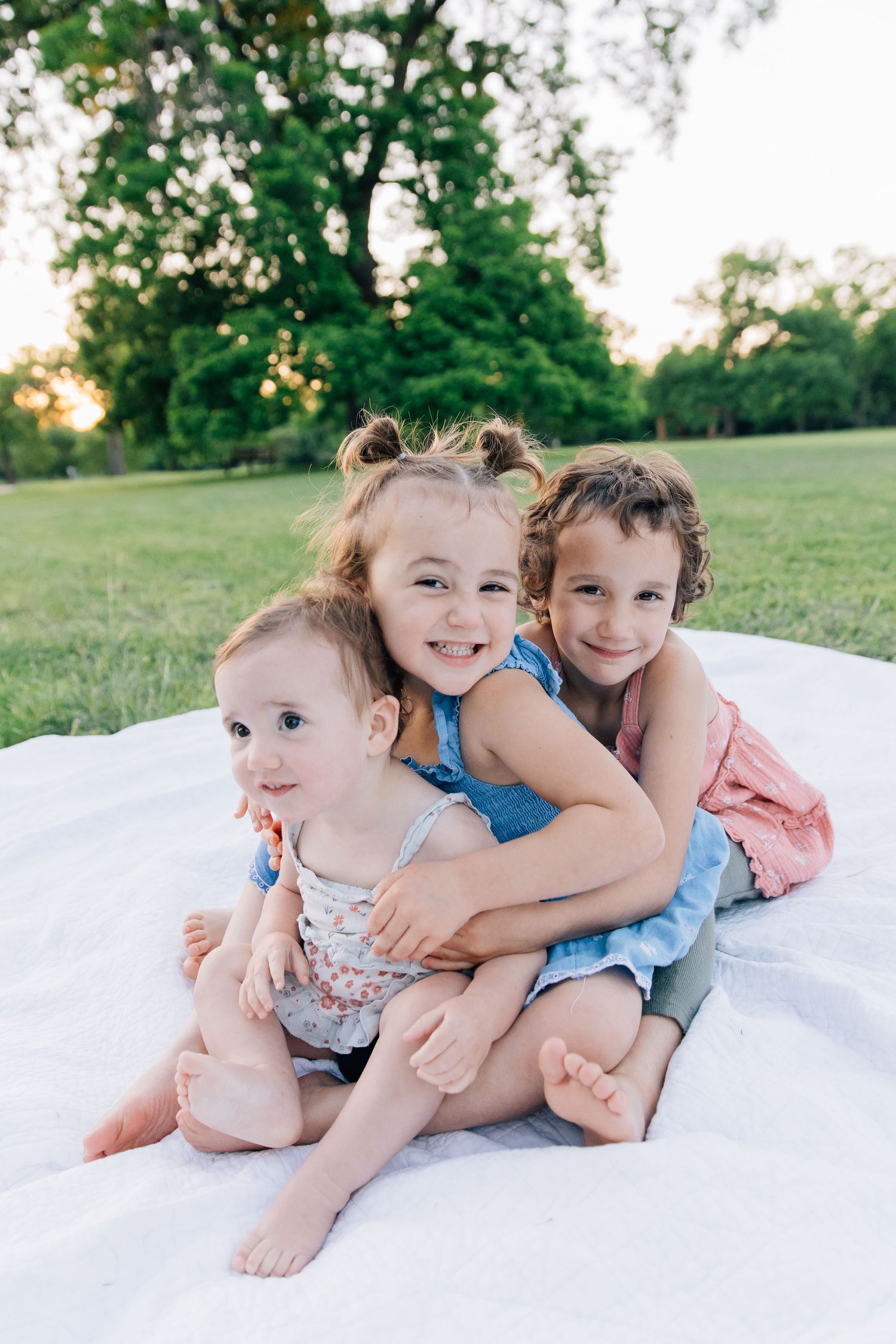 Three young children hugging and smiling on white blanket in sunny green park at golden hour.