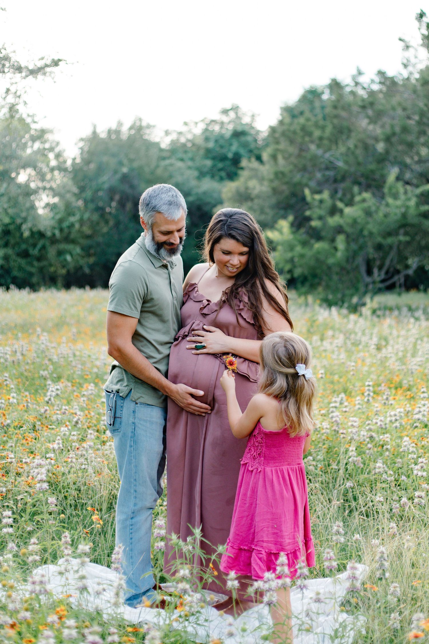 Pregnant woman in mauve dress with husband and daughter in flower field during maternity photo session.
