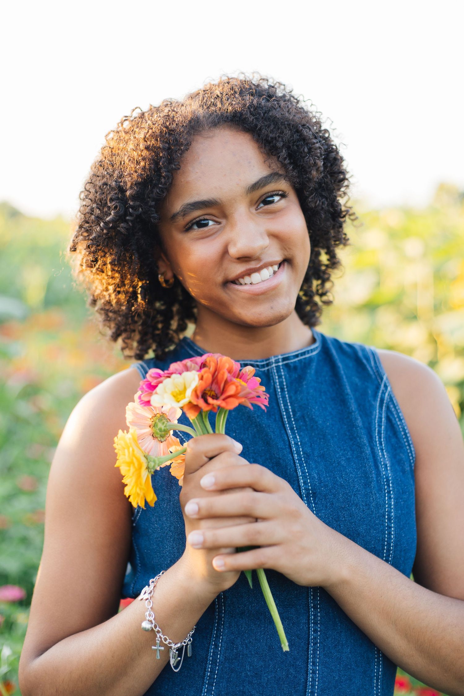 Smiling young woman with curly hair holding colorful flowers in a sunny garden, wearing a denim top and silver bracelet.