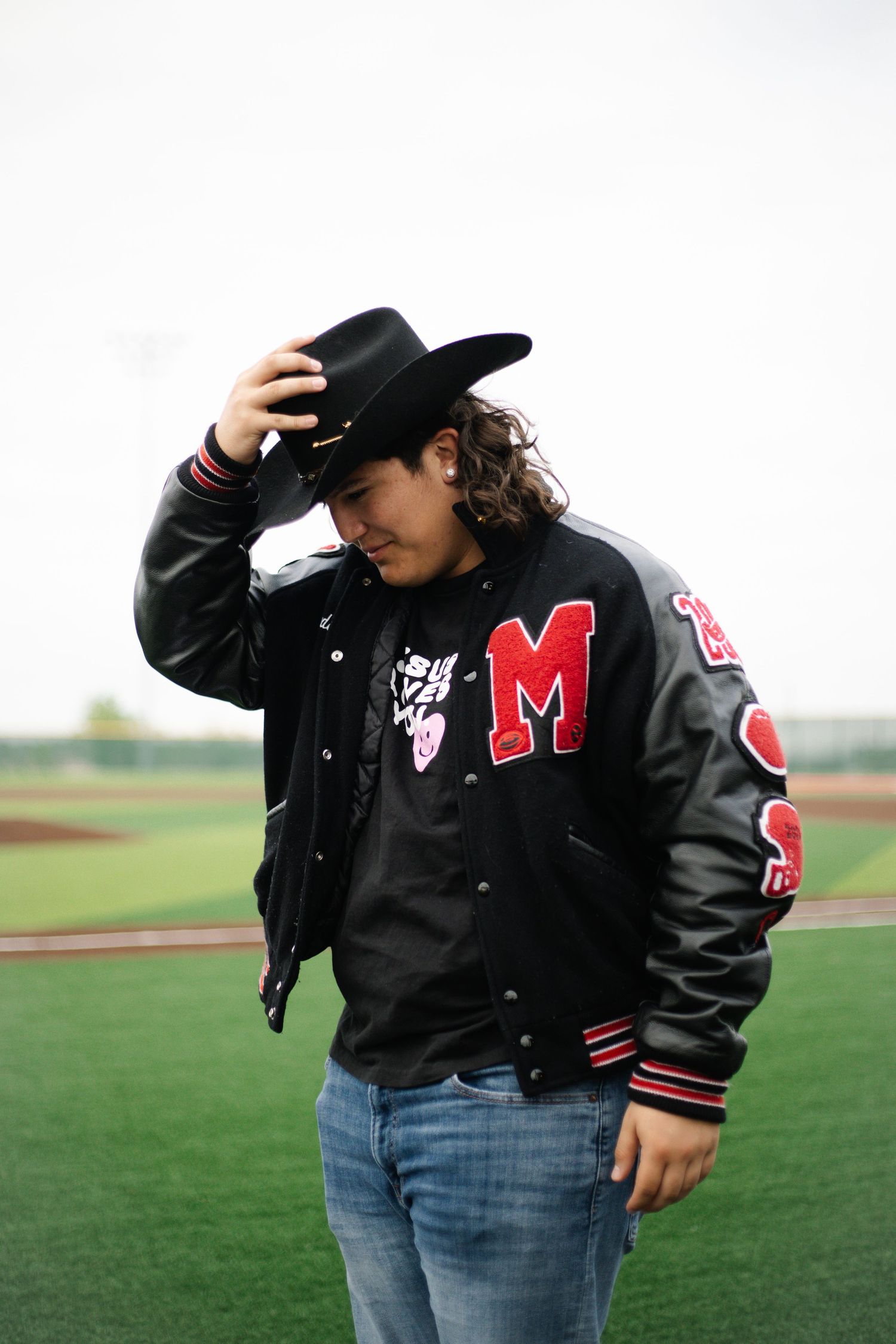 Young man in black varsity jacket with M letter patch and cowboy hat standing on baseball field.