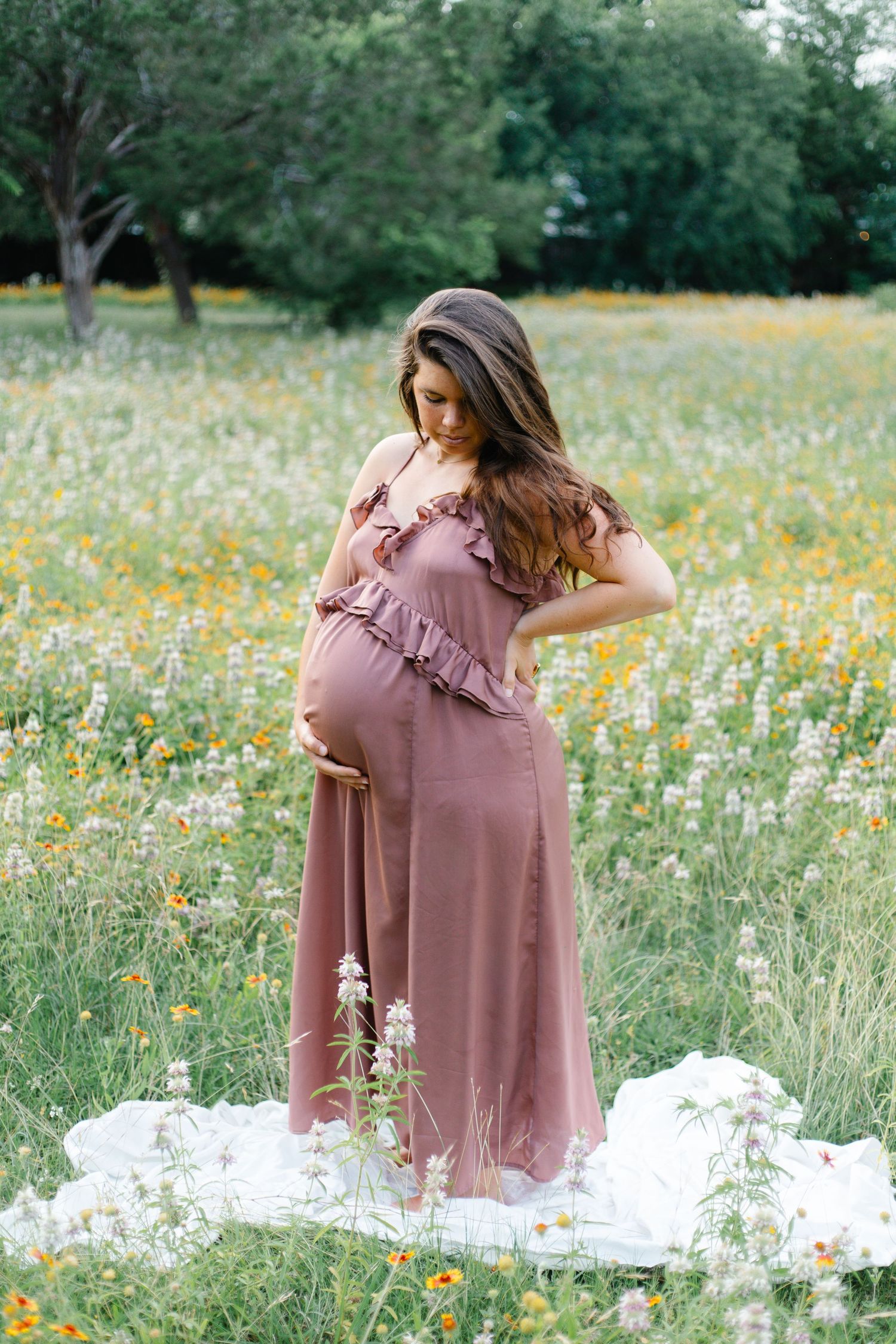Pregnant woman in mauve ruffled maxi dress standing in colorful wildflower meadow during outdoor maternity photoshoot.
