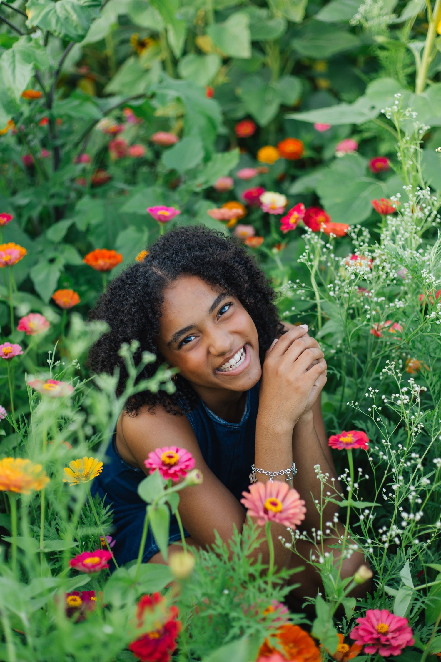 Smiling young Black woman sitting among colorful zinnia flowers in lush green garden, looking up joyfully.