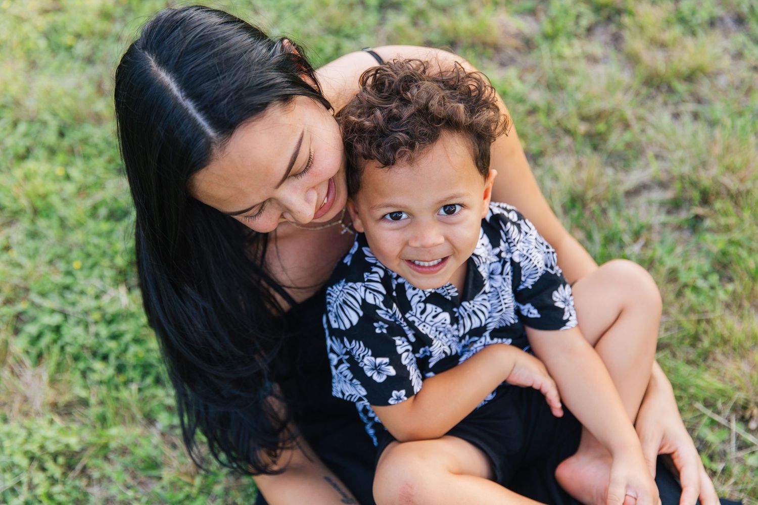 A mother with long dark hair smiles and hugs her young curly-haired boy sitting on grass outdoors.