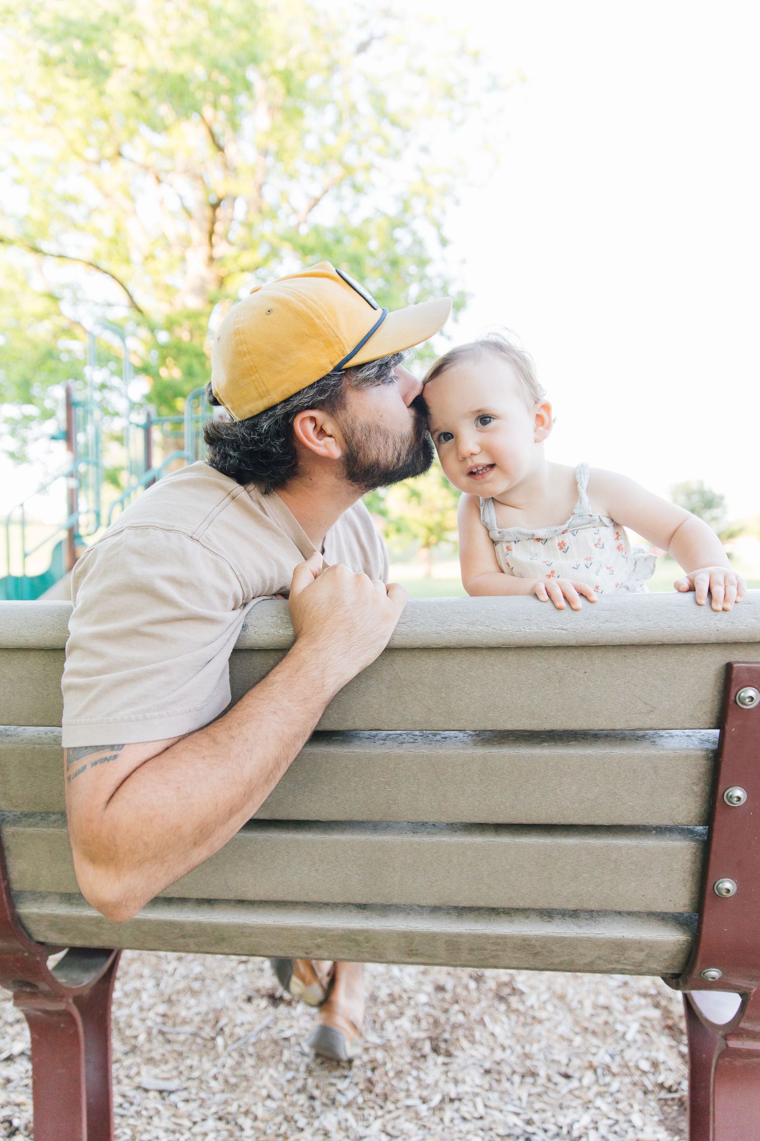 A bearded father wearing a yellow cap kisses his toddler daughter on a park bench outdoors.