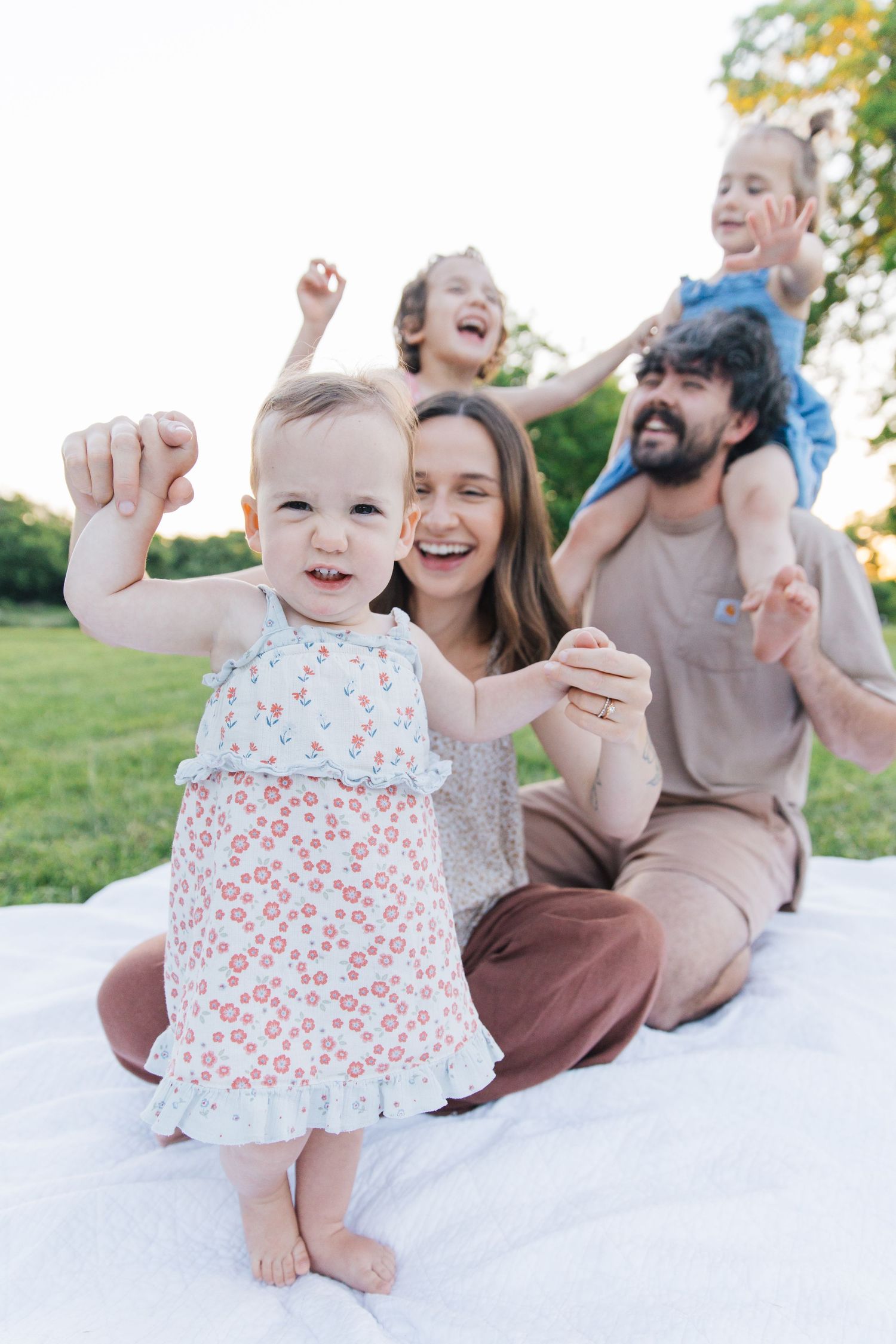 Happy family enjoying a sunny outdoor picnic on a white blanket, with parents and two young children laughing together.