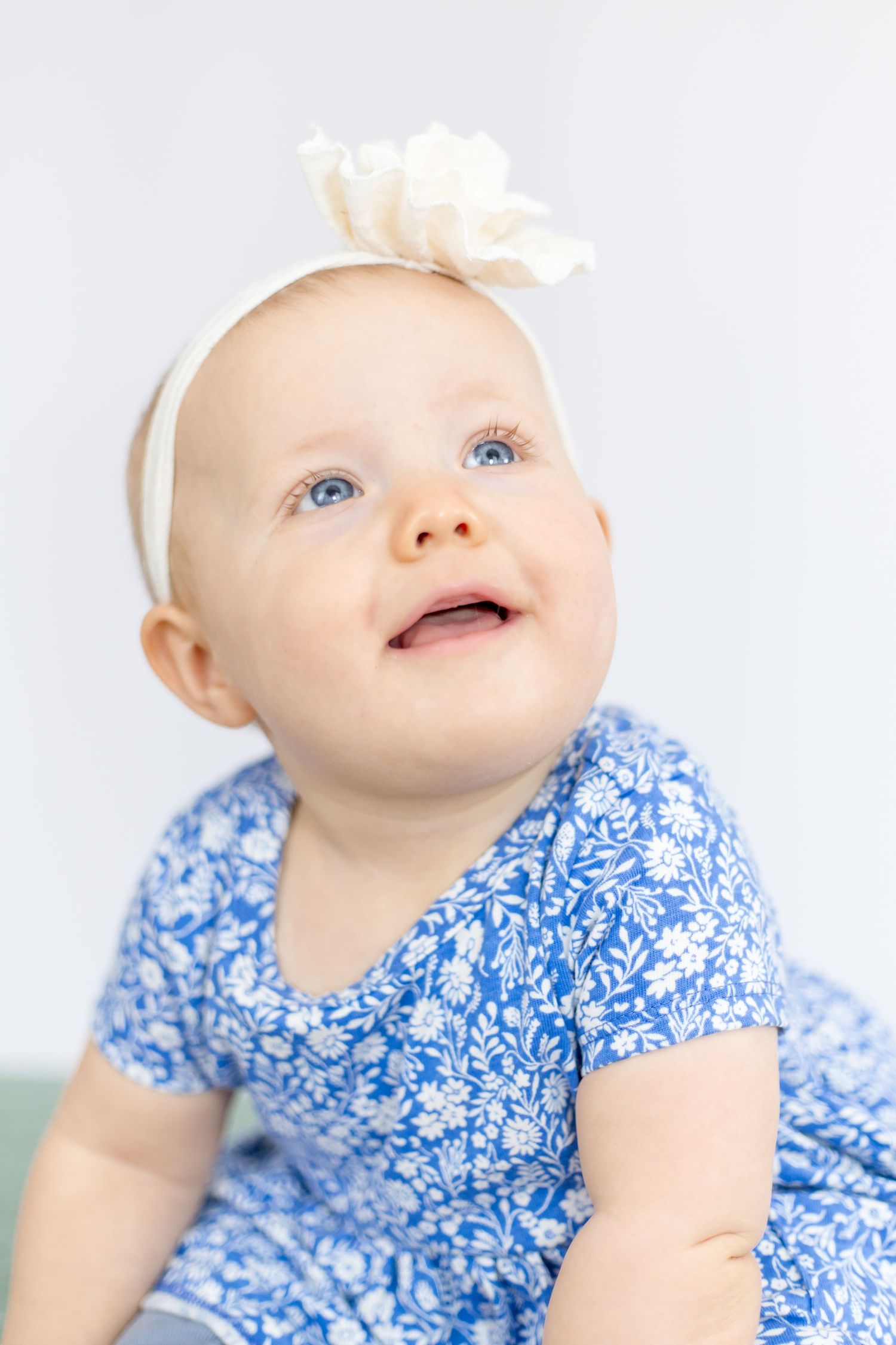 Baby with white bow headband wearing blue floral dress looking up with joyful expression.