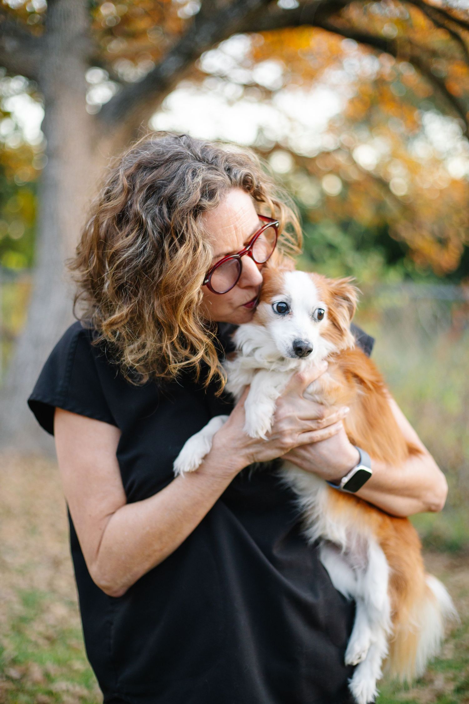 A staff member in glasses embraces a brown and white dog under autumn trees.