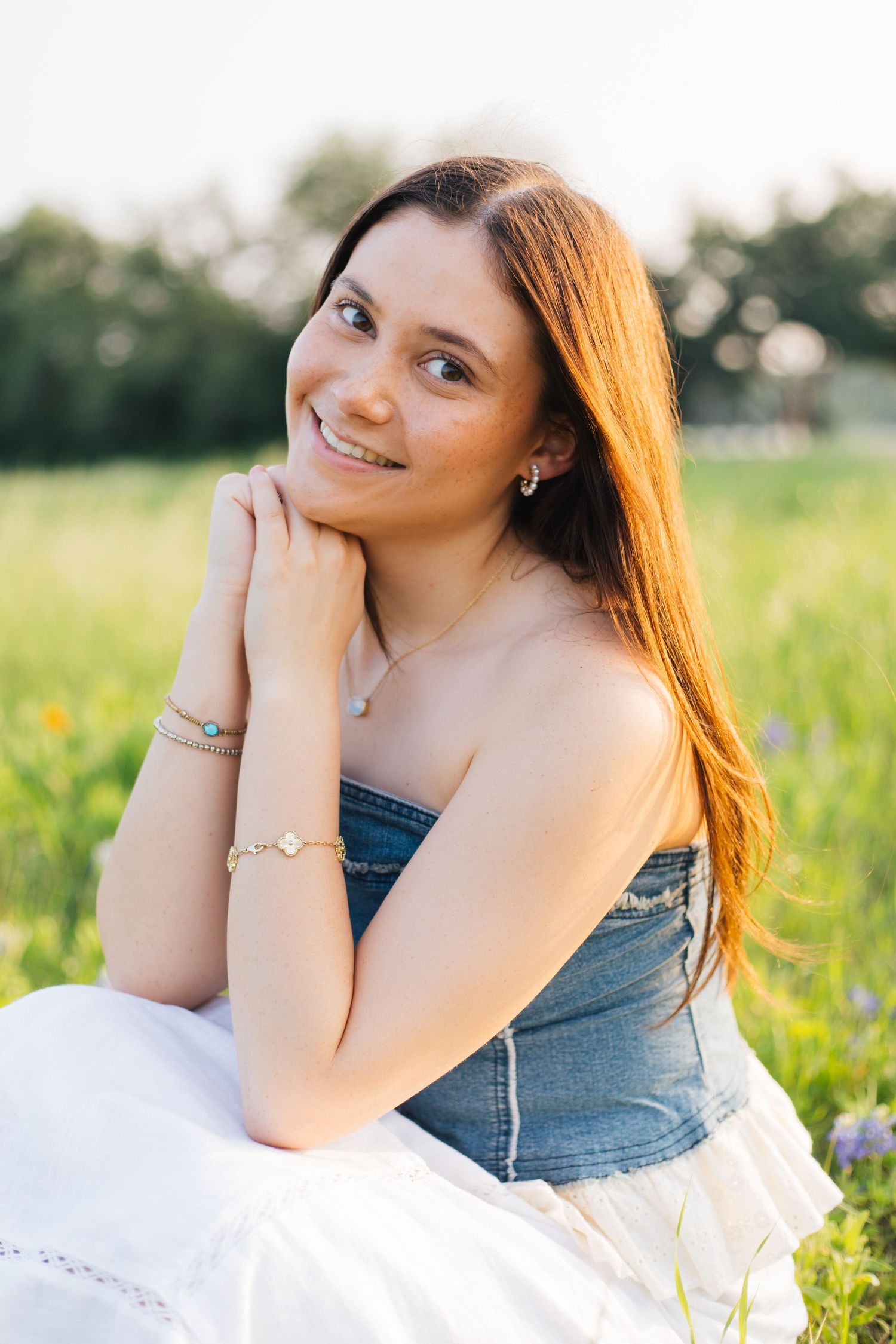 Smiling young woman with long hair in denim top and white skirt sitting in a sunny green field. Senior Portraits Round Rock TX