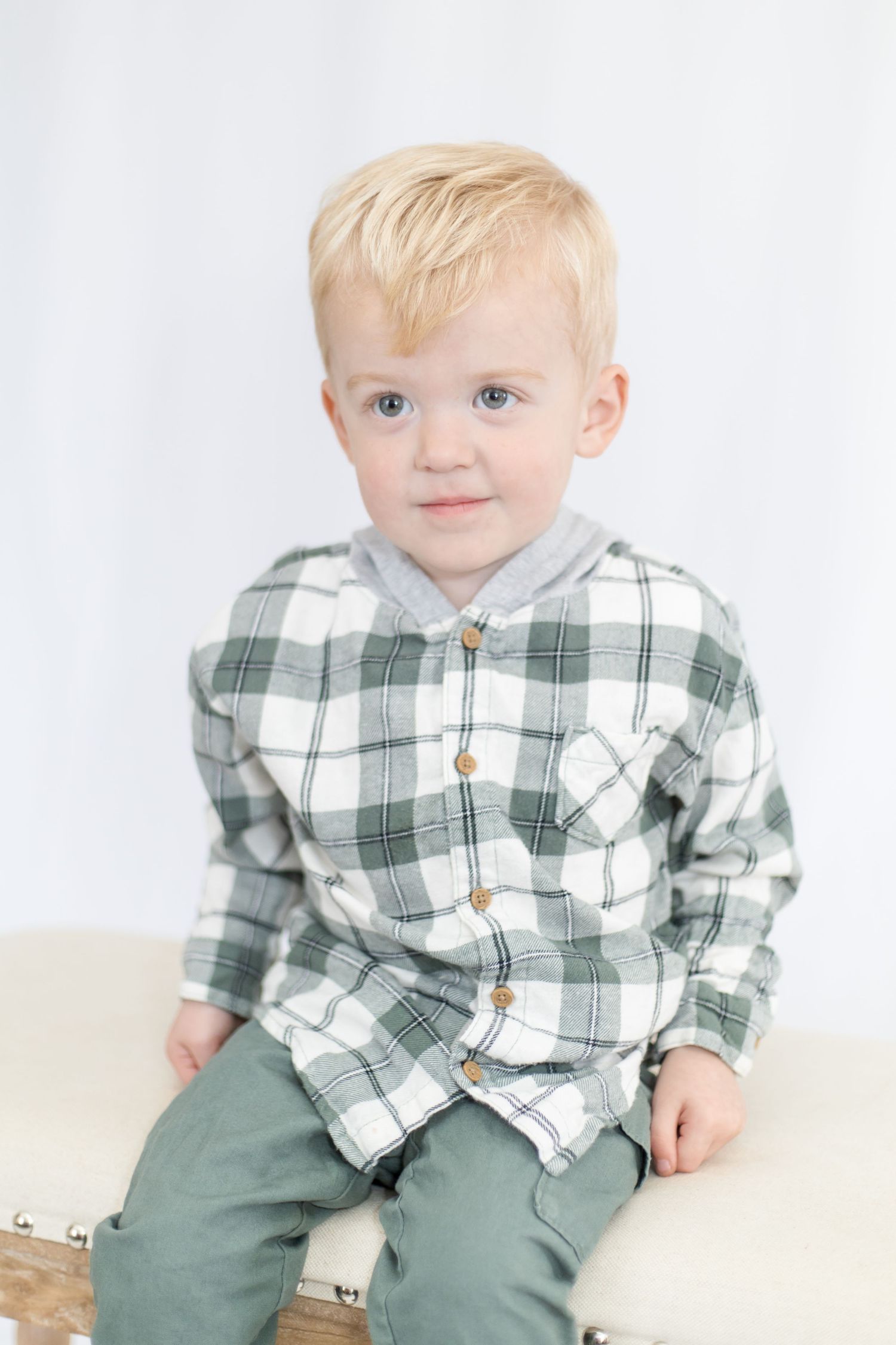 Child in plaid button-up shirt and green pants sitting against white backdrop.