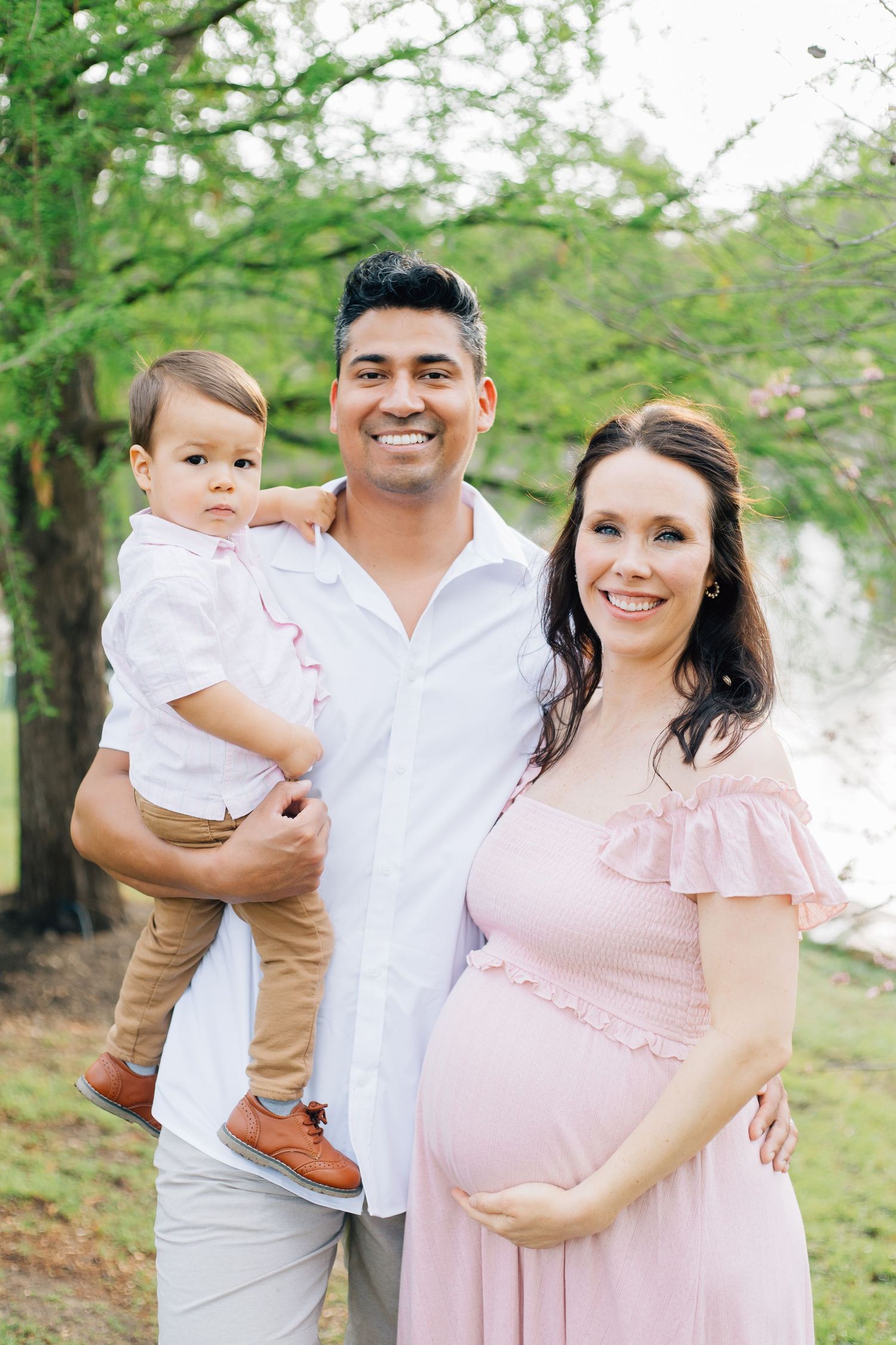 A family takes maternity photos together outdoors wearing coordinated pink and white outfits.