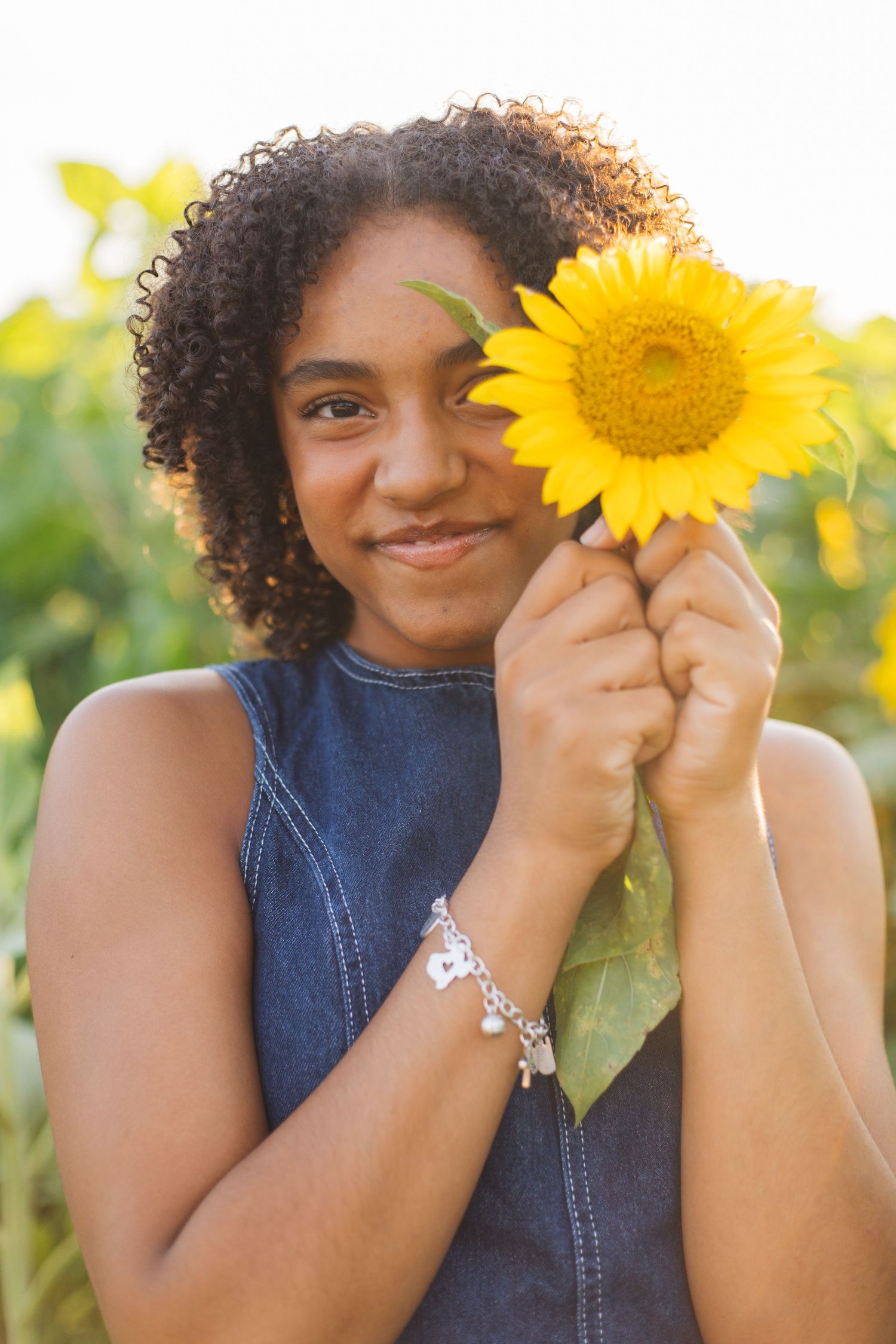 Young woman holding a bright yellow sunflower in a sunflower field, wearing a denim top and silver charm bracelet.