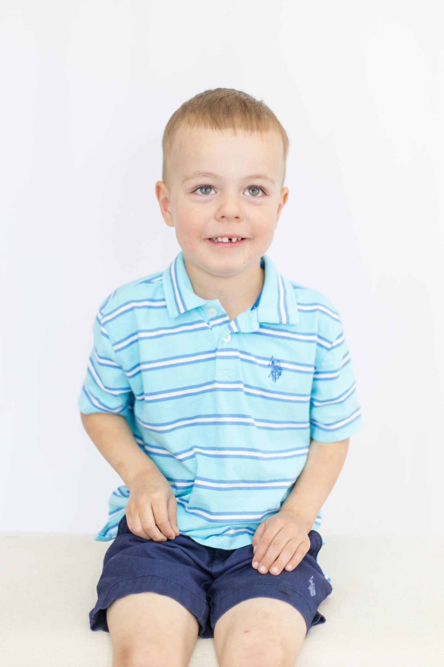 Young child in light blue striped polo shirt and navy shorts sitting against white background.