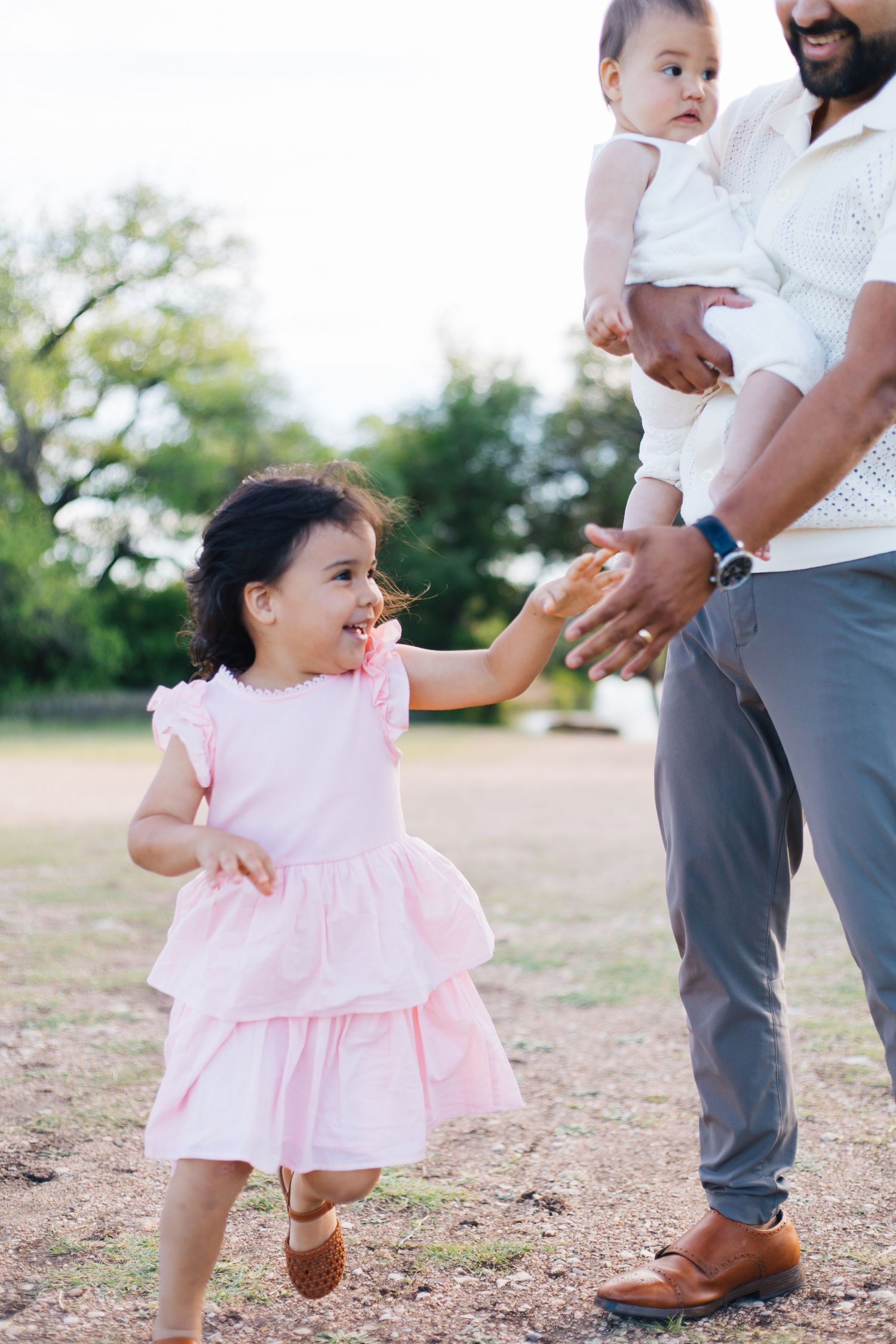 A young girl in a pink dress runs toward her father, who holds a baby outdoors in a park.