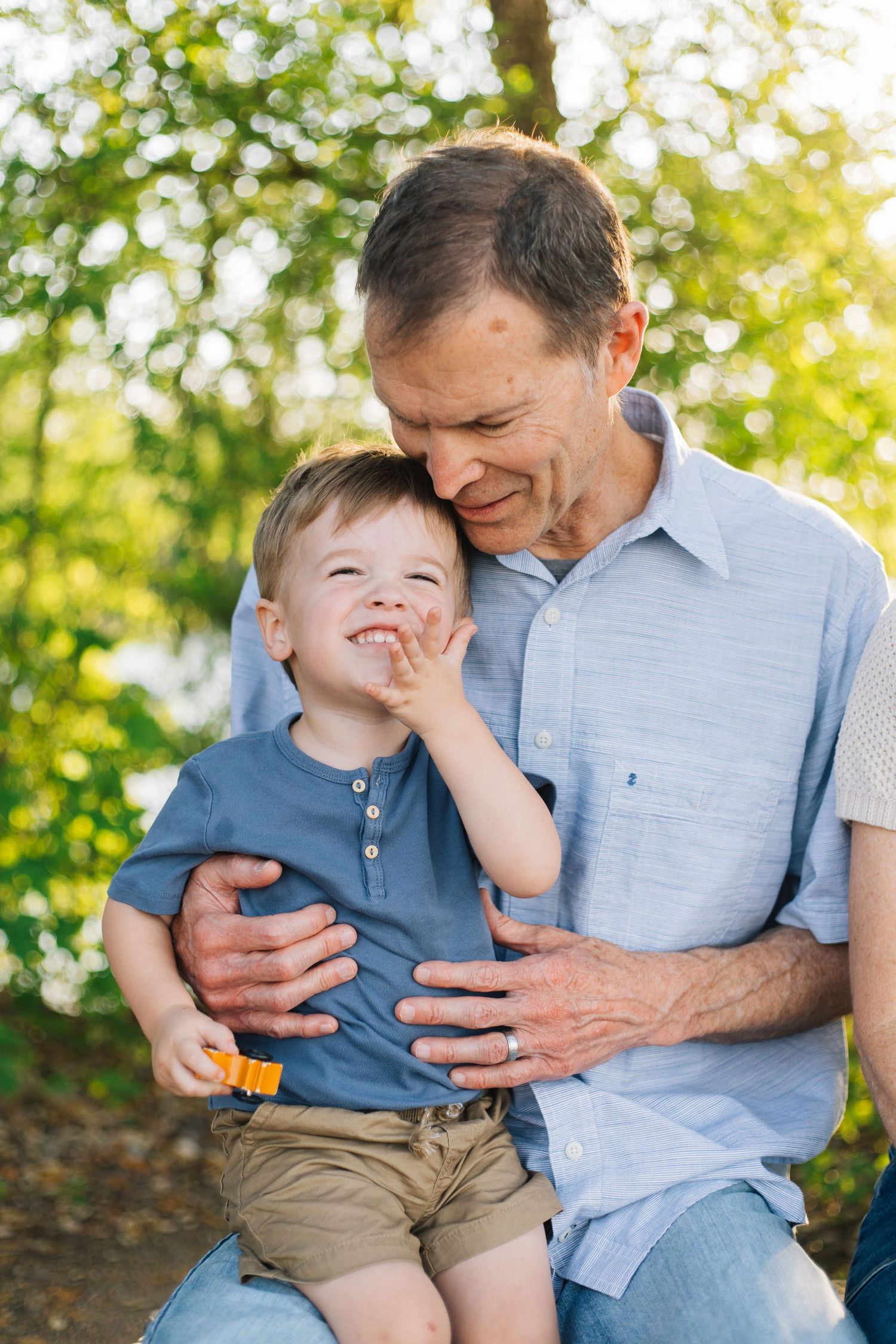 Older man smiling and hugging a laughing toddler boy outdoors in a sunny green park.
