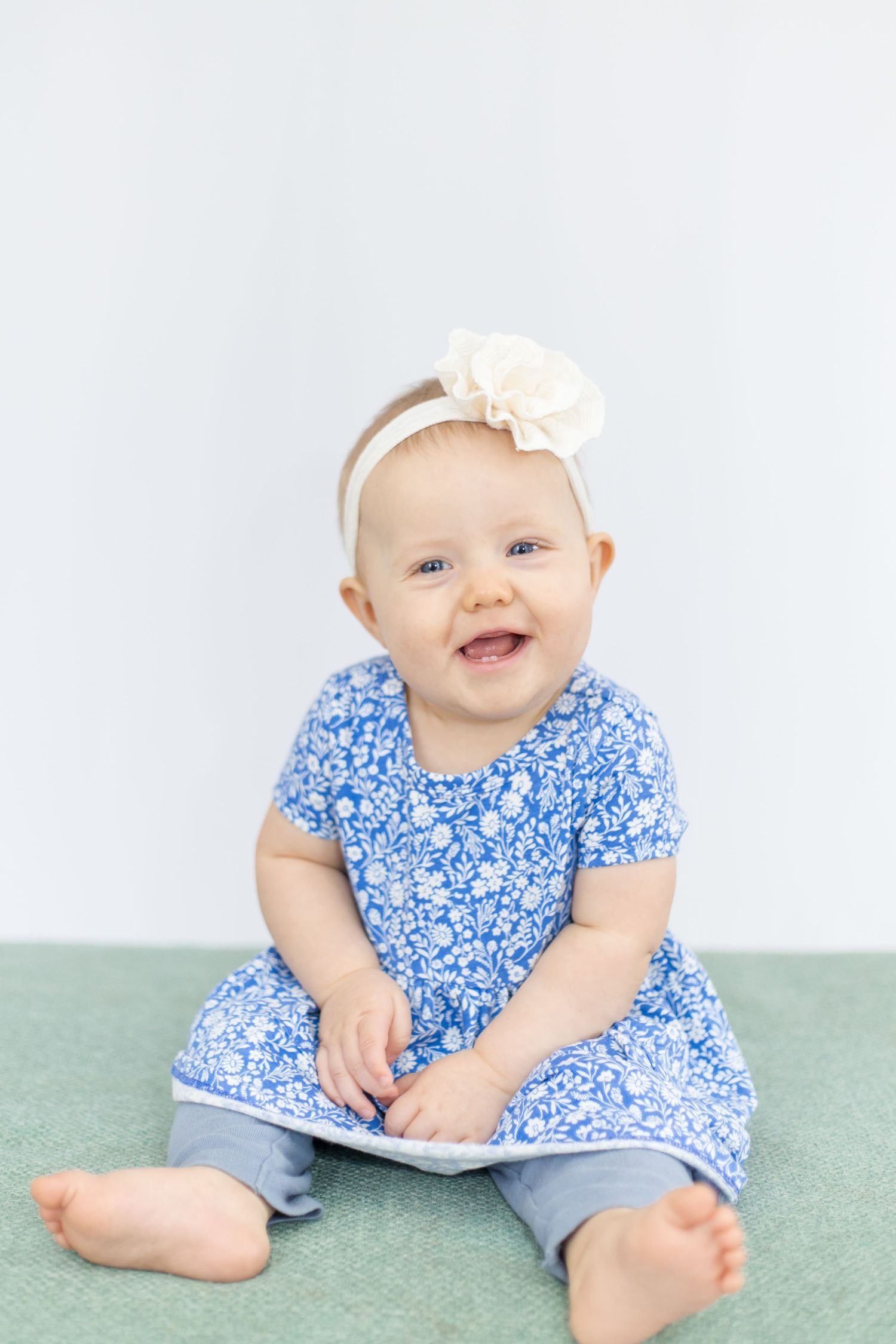 Baby in blue floral dress and white headband sitting on mint green surface.
