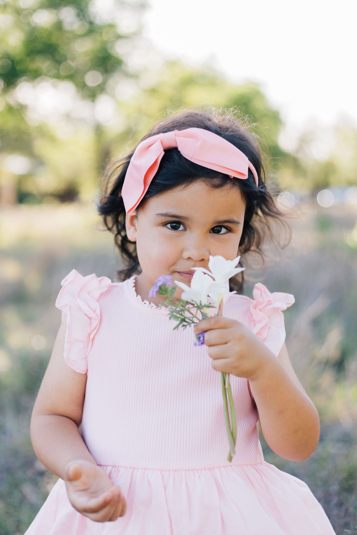 Young girl in pink dress and bow headband holds white wildflowers in an outdoor garden setting.