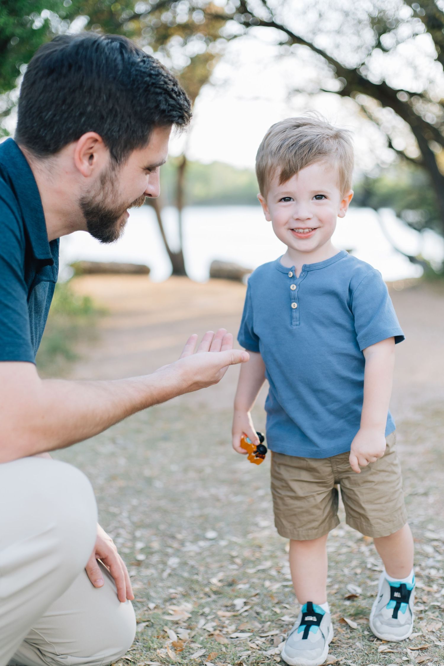 Father crouching down to interact with his smiling toddler son in a park near a lake.