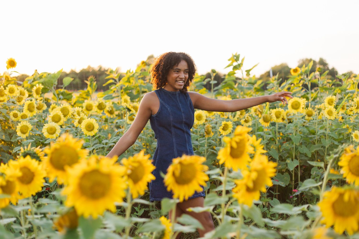 A joyful woman in a blue dress smiles while walking through a vibrant sunflower field in golden sunlight.