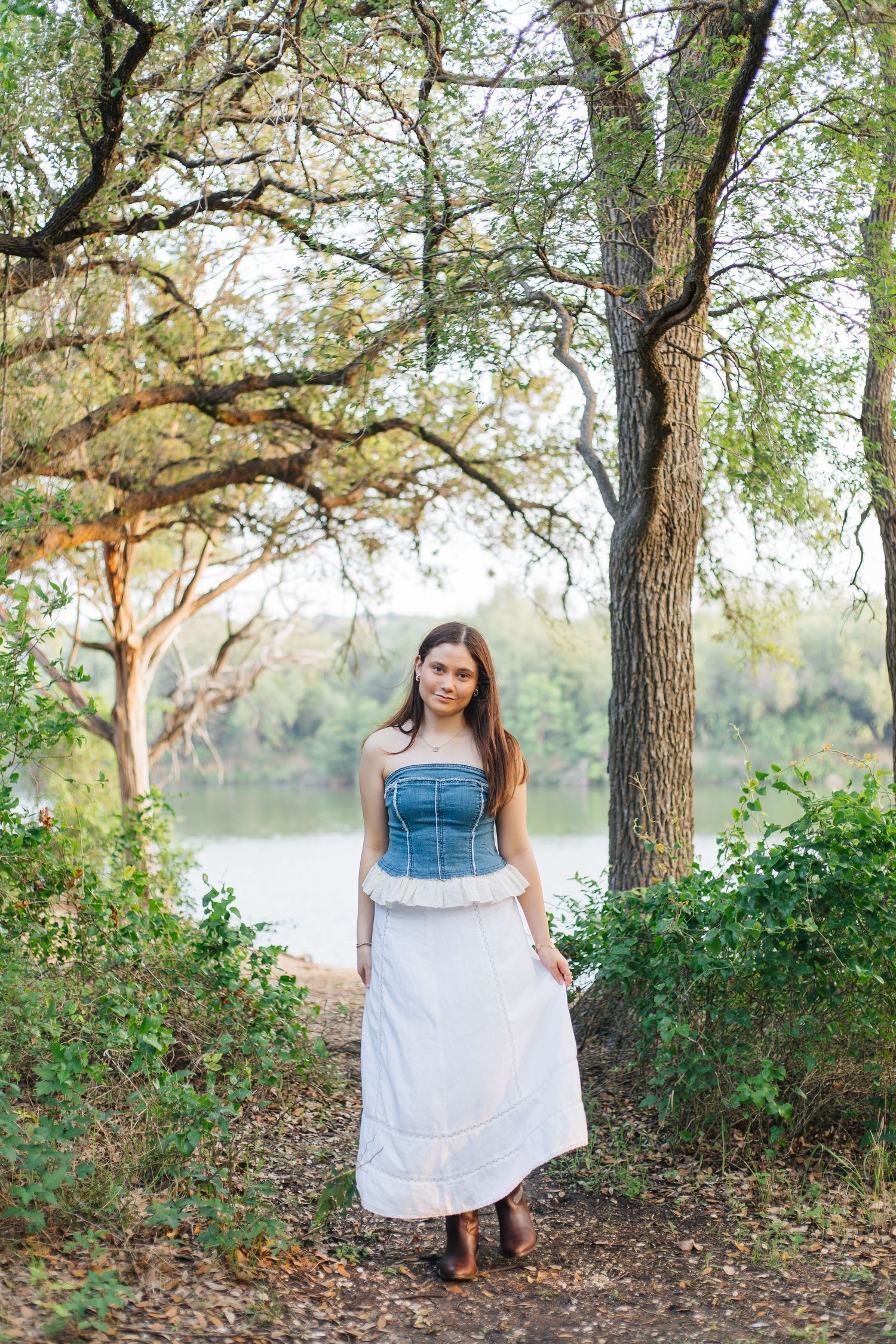 Young woman in white skirt and denim corset top standing among trees near a serene river in natural outdoor setting. Senior Portraits